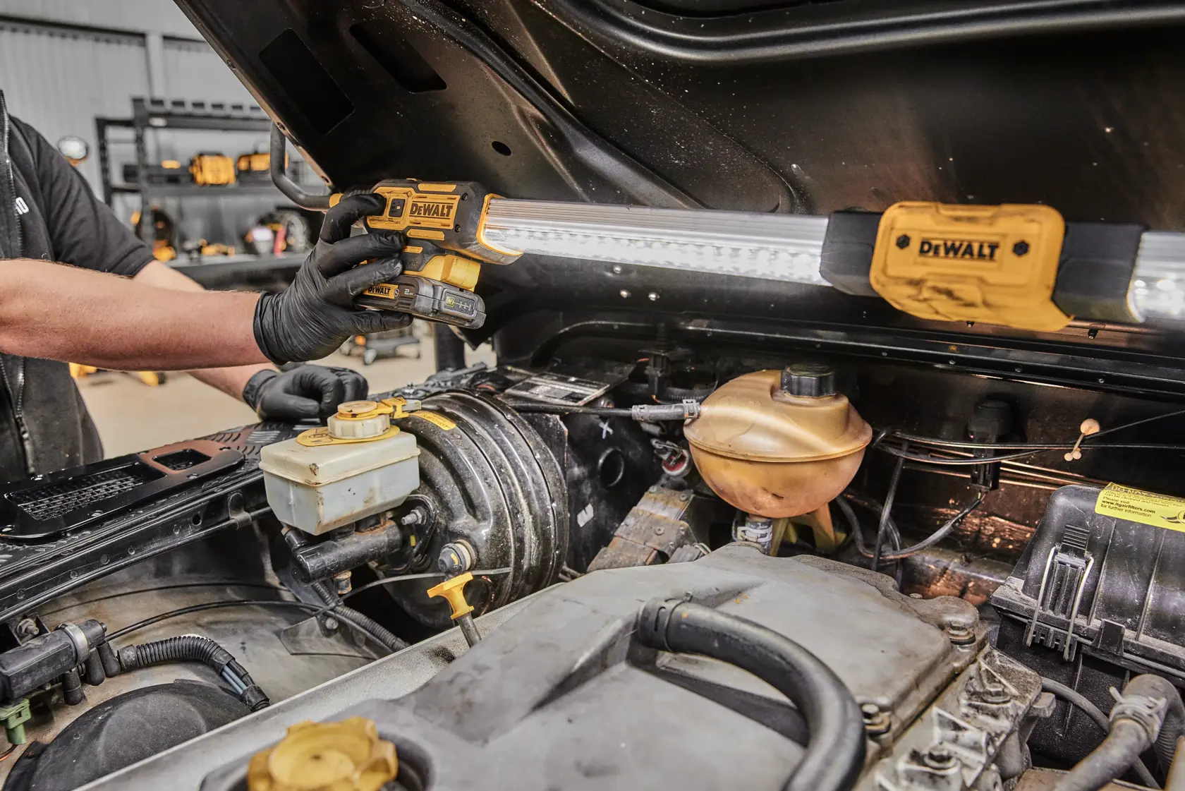 Un mecánico utiliza una lámpara de trabajo DEWALT DCL045 para iluminar el compartimento del motor de un vehículo. La lámpara está montada bajo el capó abierto para proporcionar una luz brillante durante las tareas de mantenimiento y reparación.