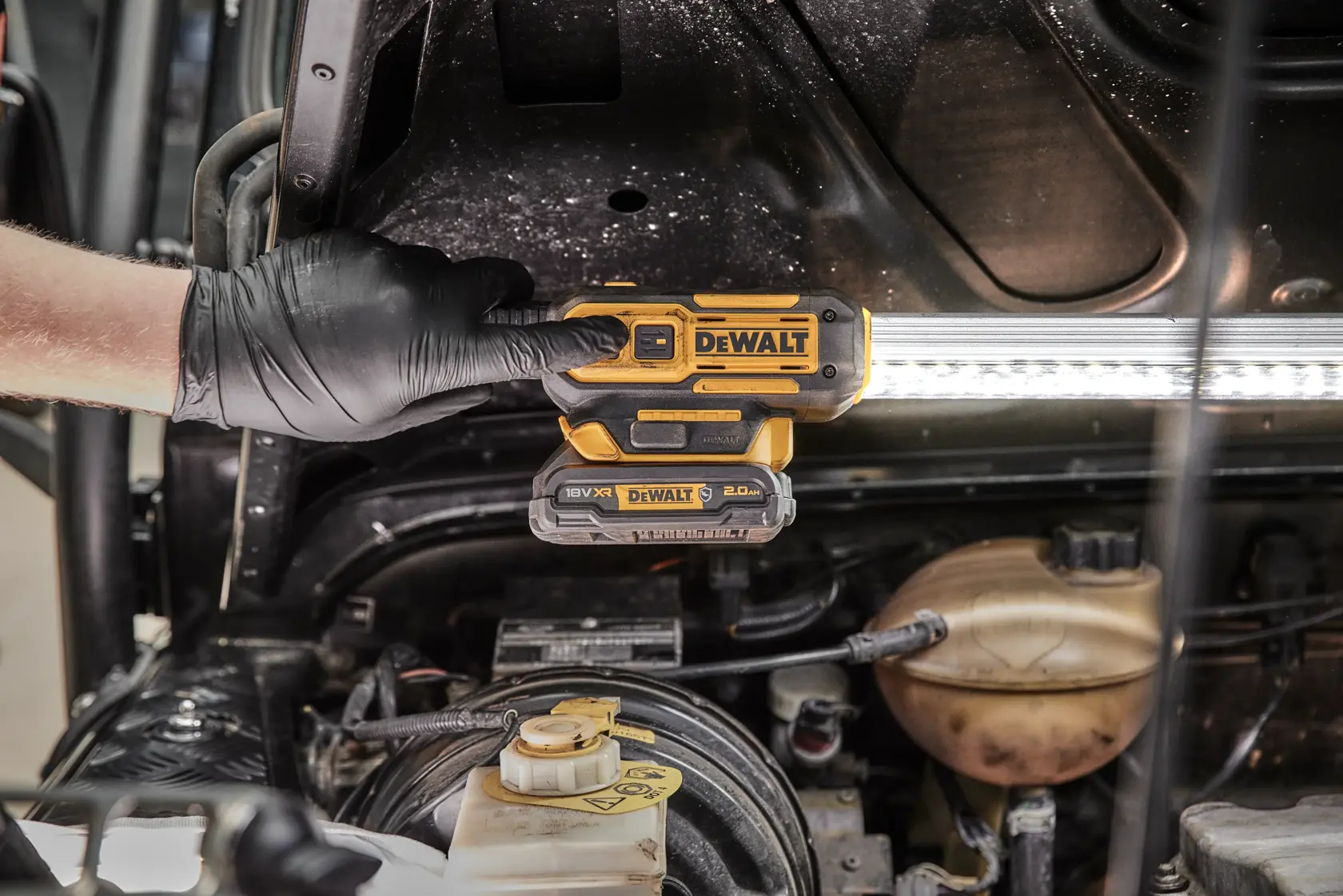 Una mano enguantada sostiene una luz de trabajo LED inalámbrica DEWALT DCL045 bajo el capó de un vehículo, iluminando el área del motor.