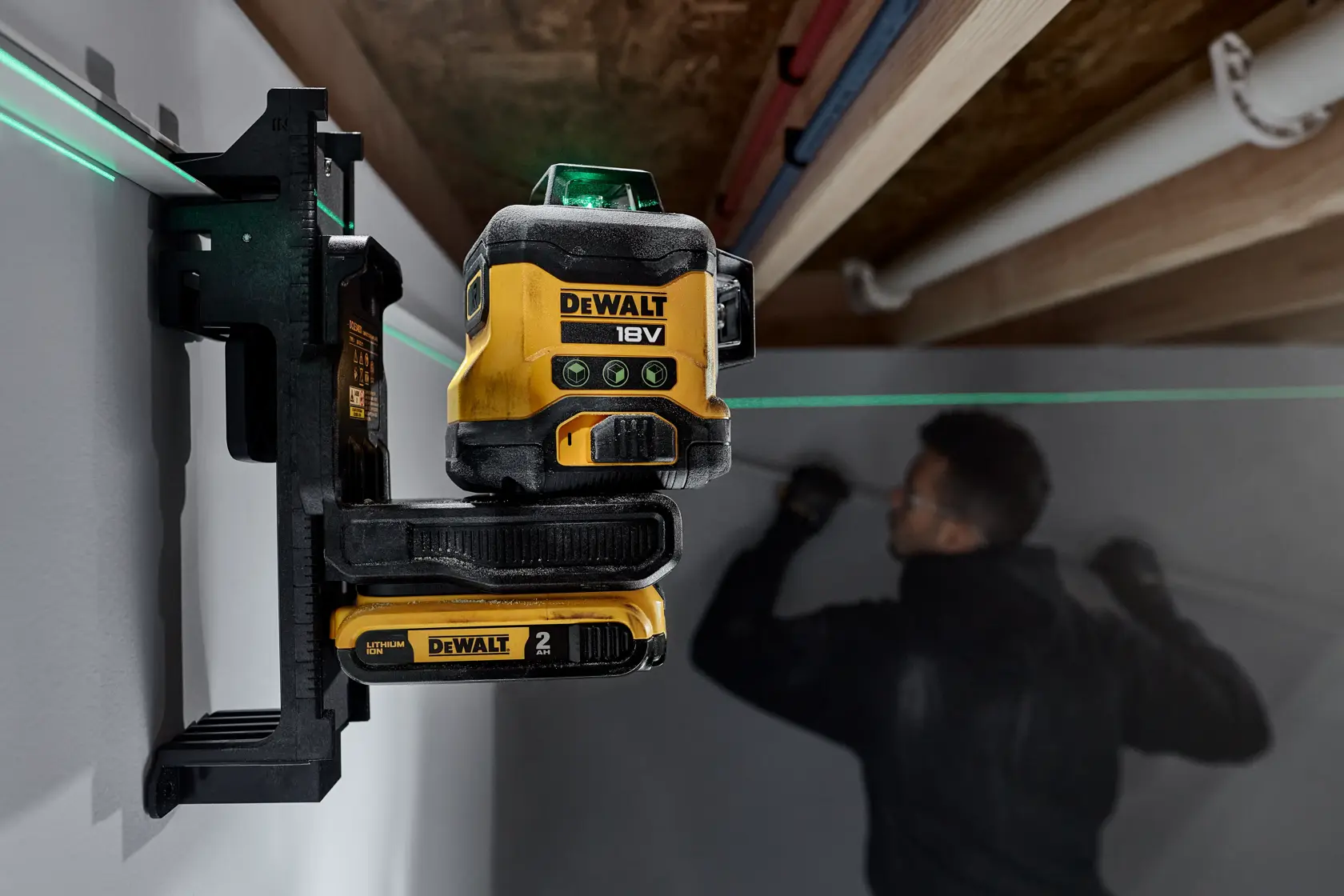 Nivel láser de líneas cruzadas DEWALT 18V montado en una pared, proyectando una línea láser verde a través de una habitación donde una persona trabaja en la pared al fondo.