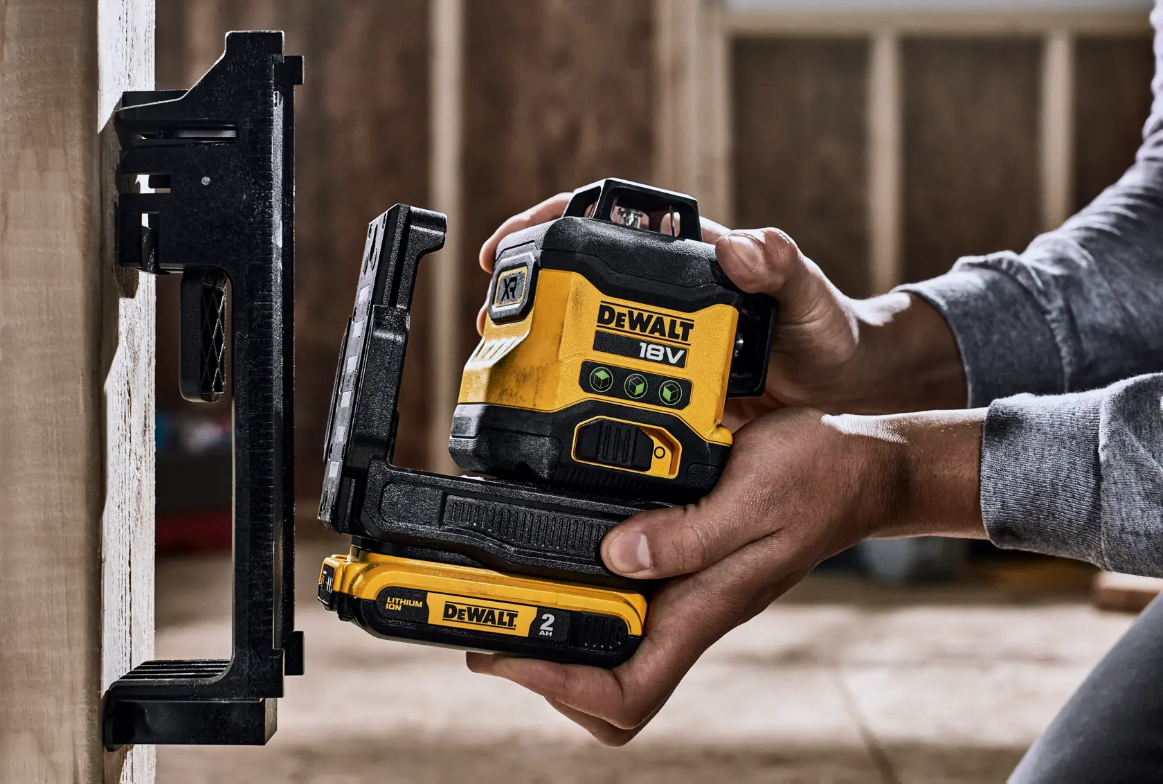 Una persona sostiene un nivel láser DEWALT de 18V con una batería de 2 Ah y lo coloca junto a un soporte montado en la pared en un entorno de construcción interior.