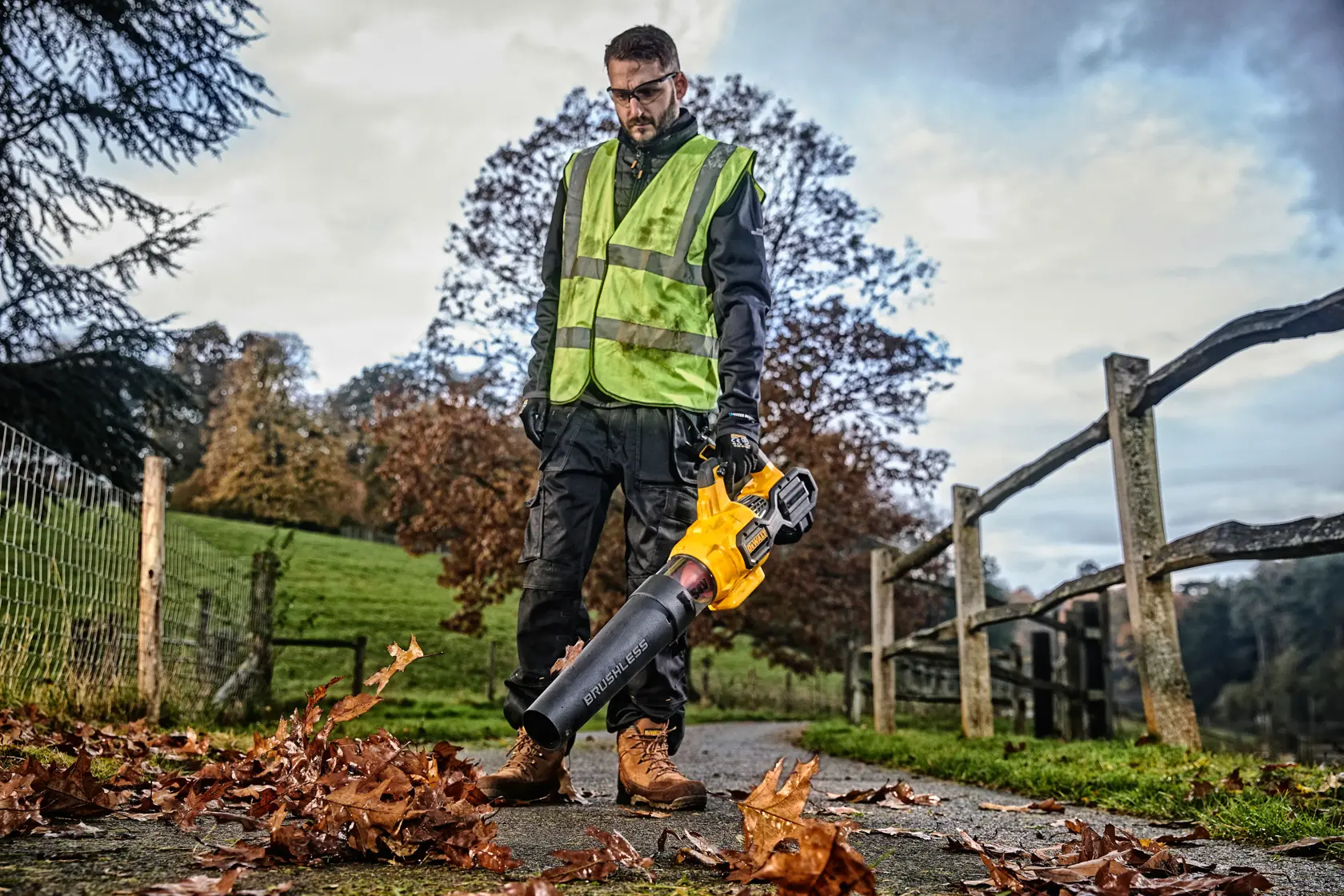Persona usando soplador de hojas DEWALT al aire libre, con chaleco de seguridad y guantes, limpiando hojas del sendero.