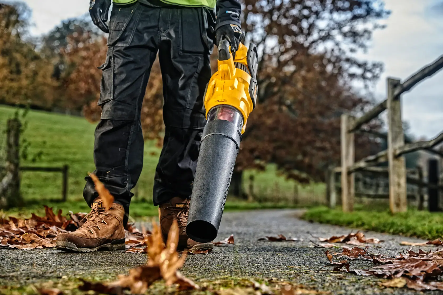 Persona usa soplador de hojas DEWALT amarillo en un camino con hojas esparcidas al aire libre.