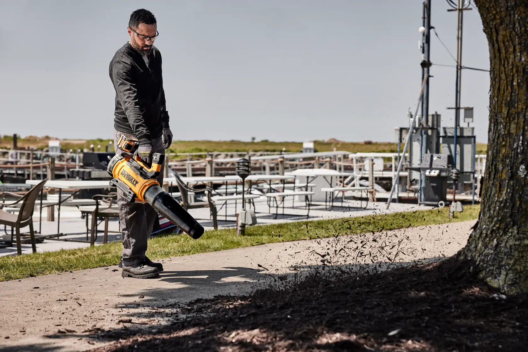 Person uses a DEWALT cordless leaf blower outdoors to clear debris from a sidewalk near tables and a tree.