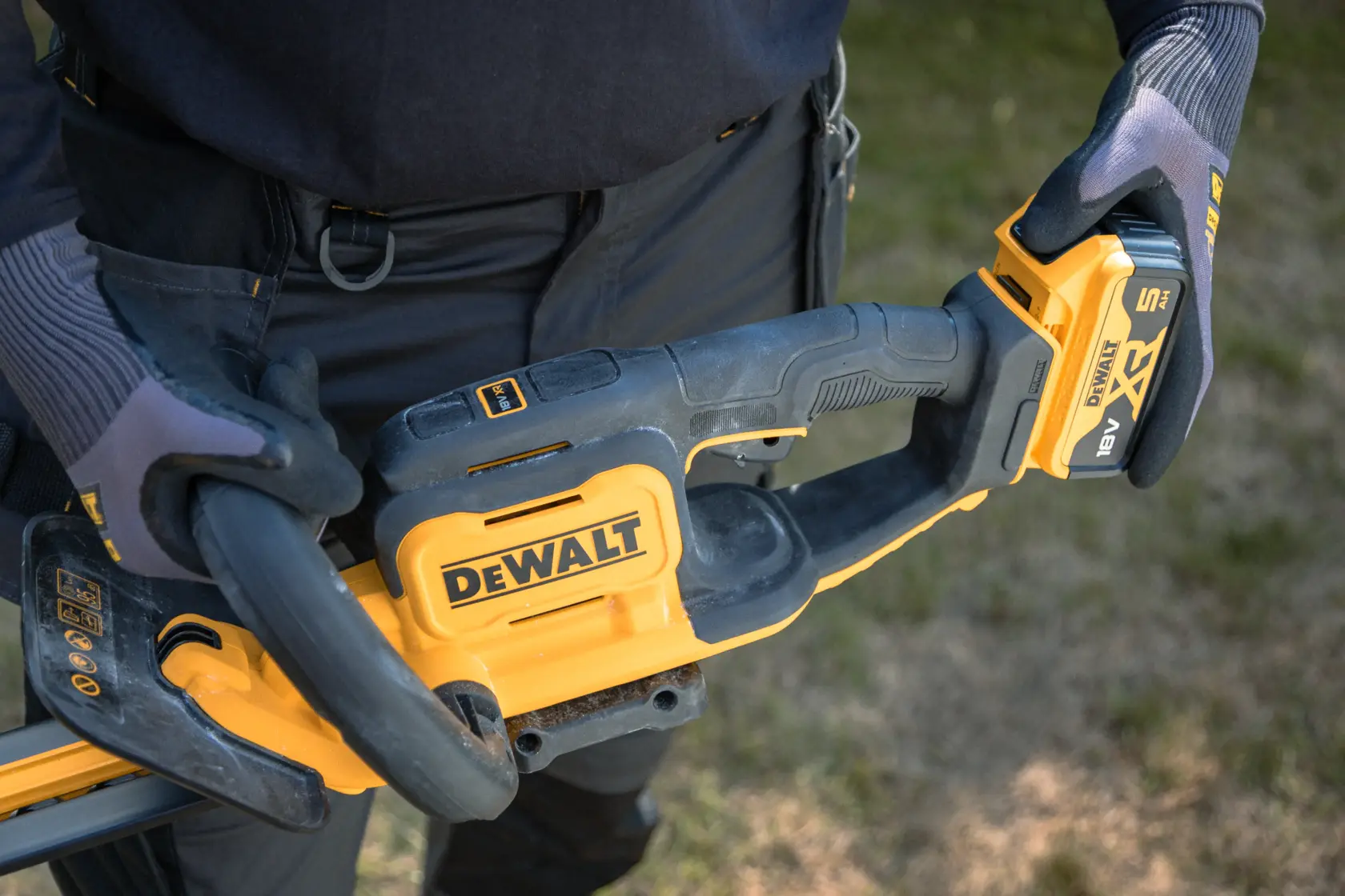 Primer plano de guantes sosteniendo un cortasetos DEWALT inalámbrico con batería al aire libre.