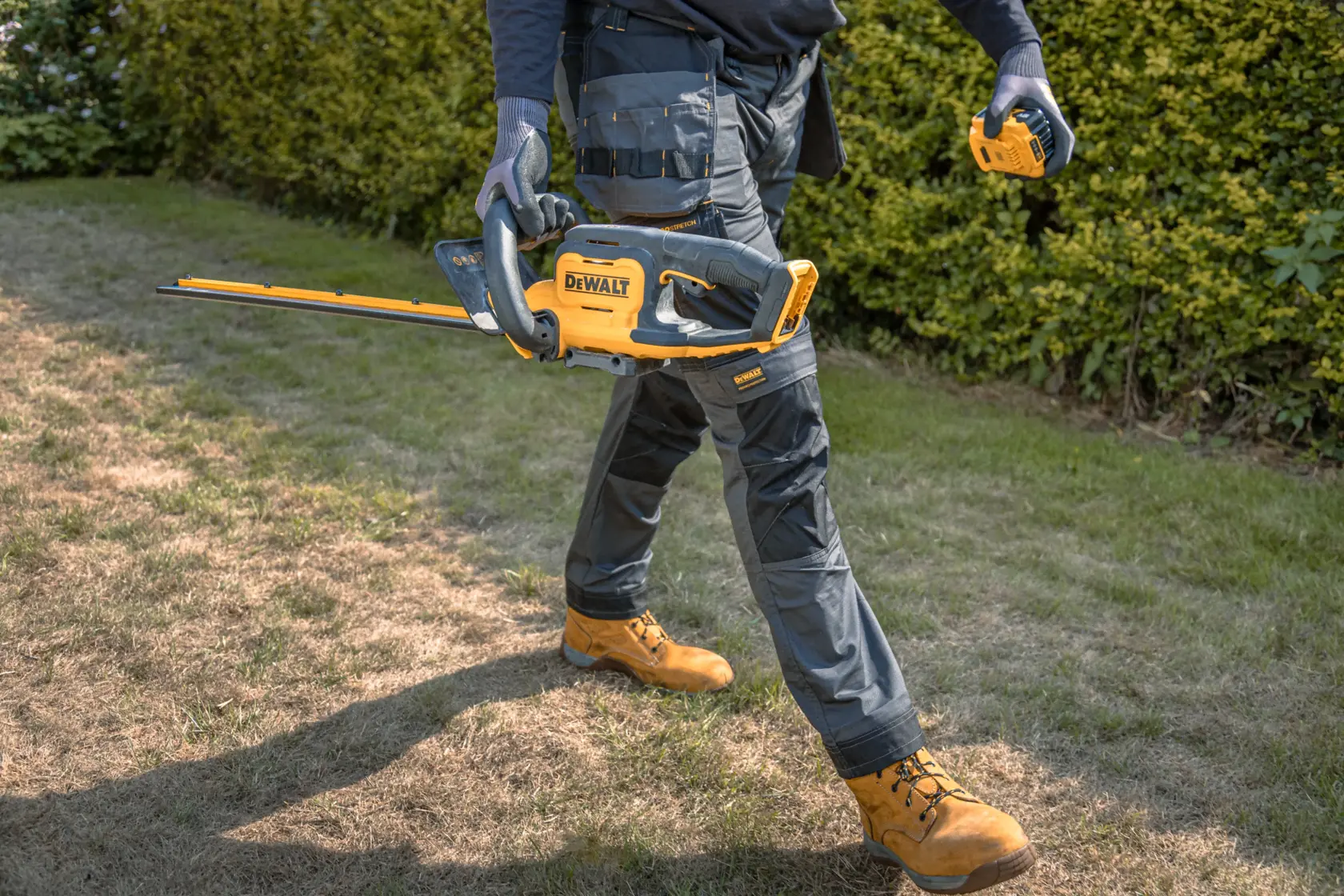Una persona con guantes de trabajo y ropa protectora sostiene un cortasetos inalámbrico DEWALT amarillo y negro y camina sobre un área de césped junto a arbustos.
