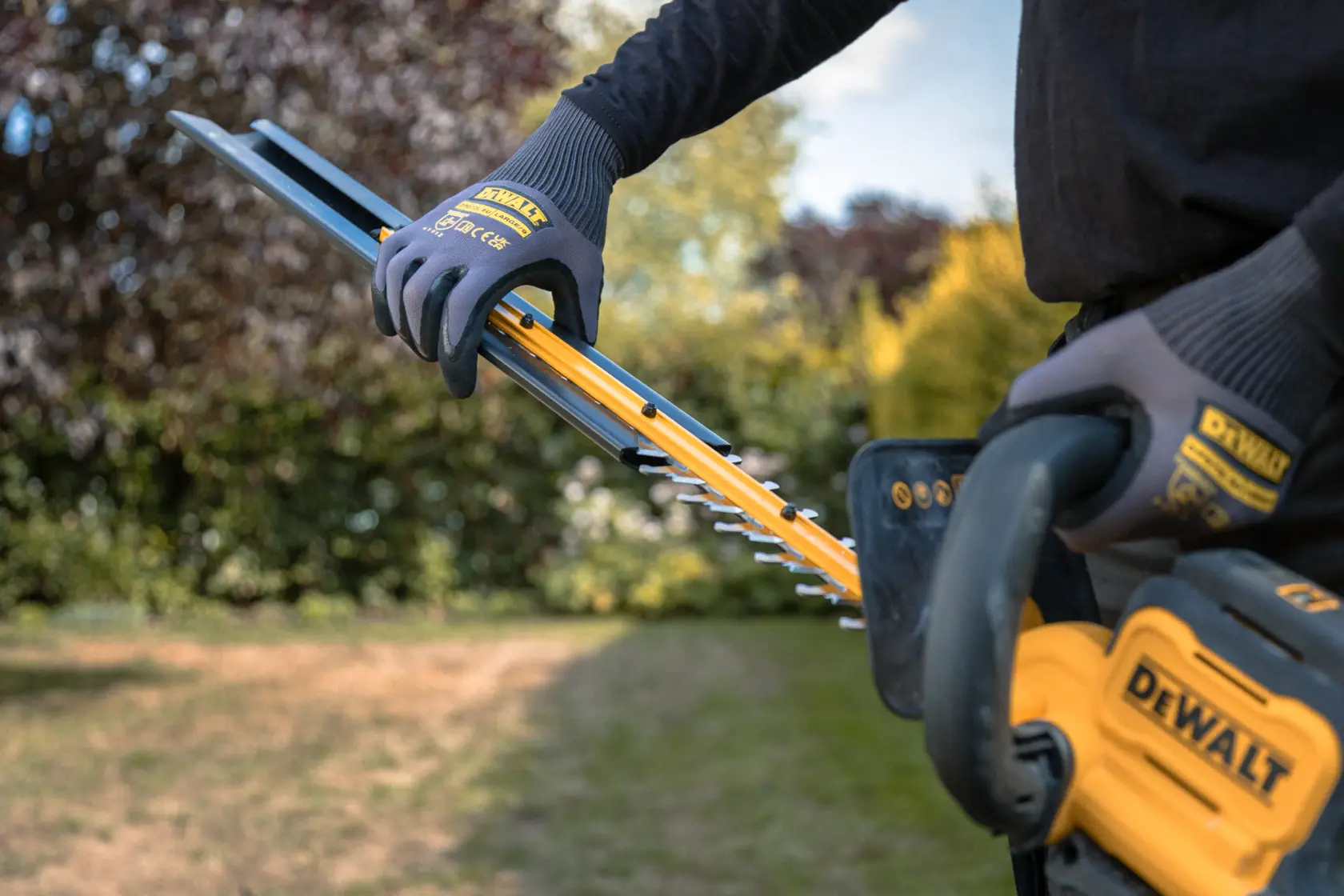 Persona usando cortasetos inalámbrico DEWALT en un jardín, con guantes protectores.
