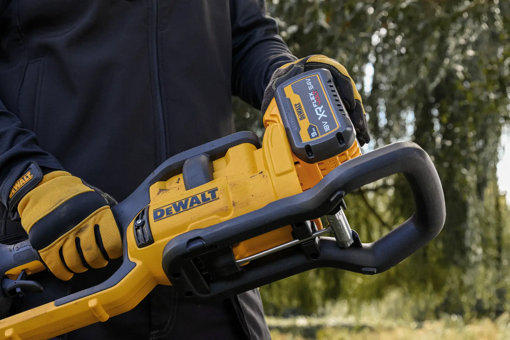 Manos con guantes sostienen motosierra DEWALT inalámbrica con batería XR FLEXVOLT 18V visible al aire libre.