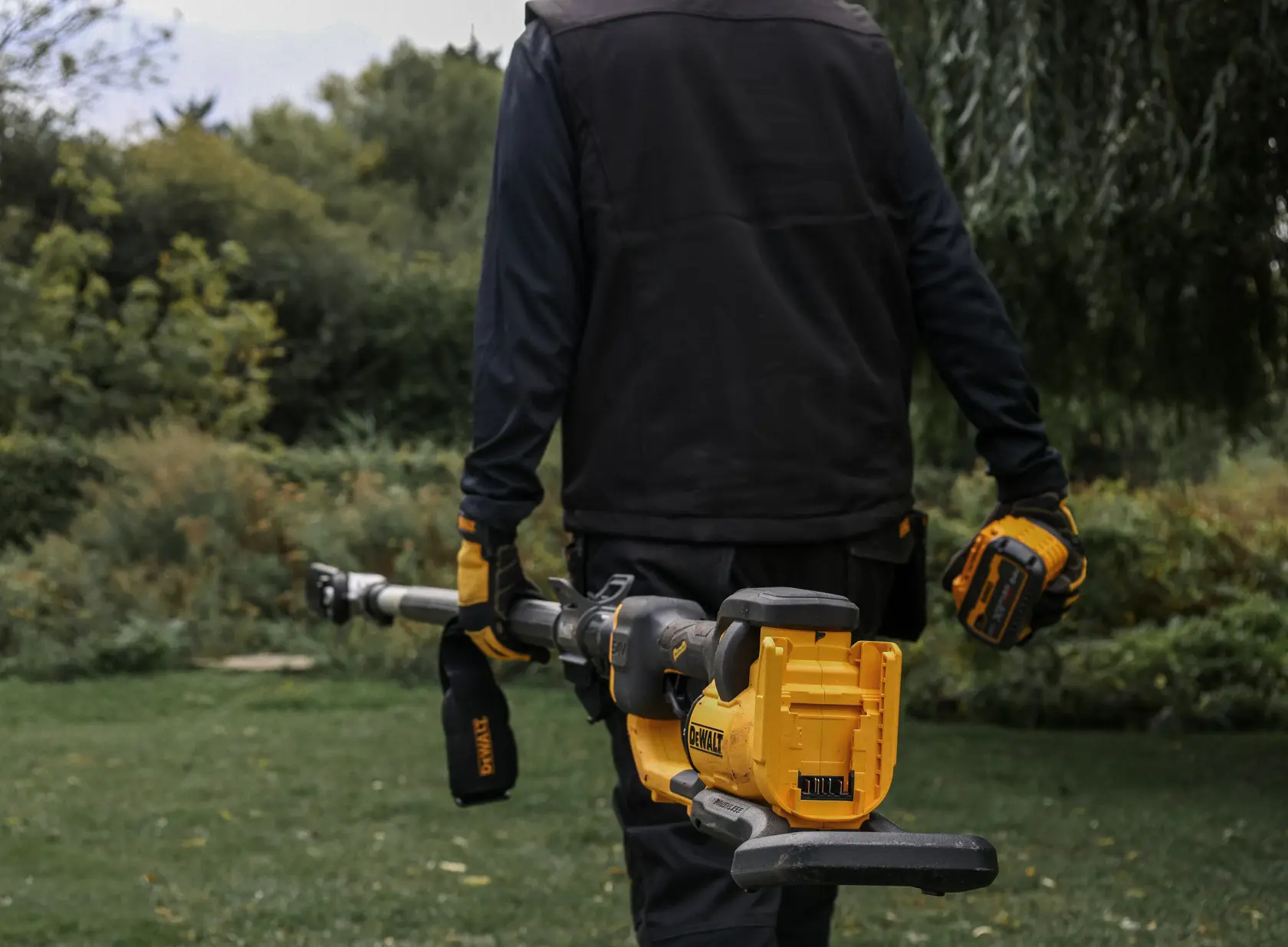 Persona sostiene podadora eléctrica DEWALT amarilla, con guantes y ropa de trabajo en un jardín.