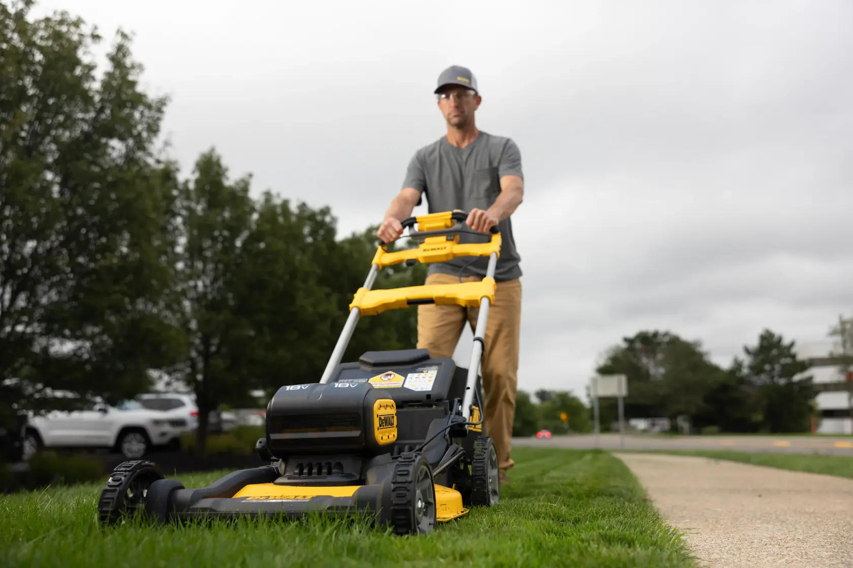 A person is using a DEWALT cordless lawn mower to cut grass along a sidewalk. The mower is black and yellow, and the