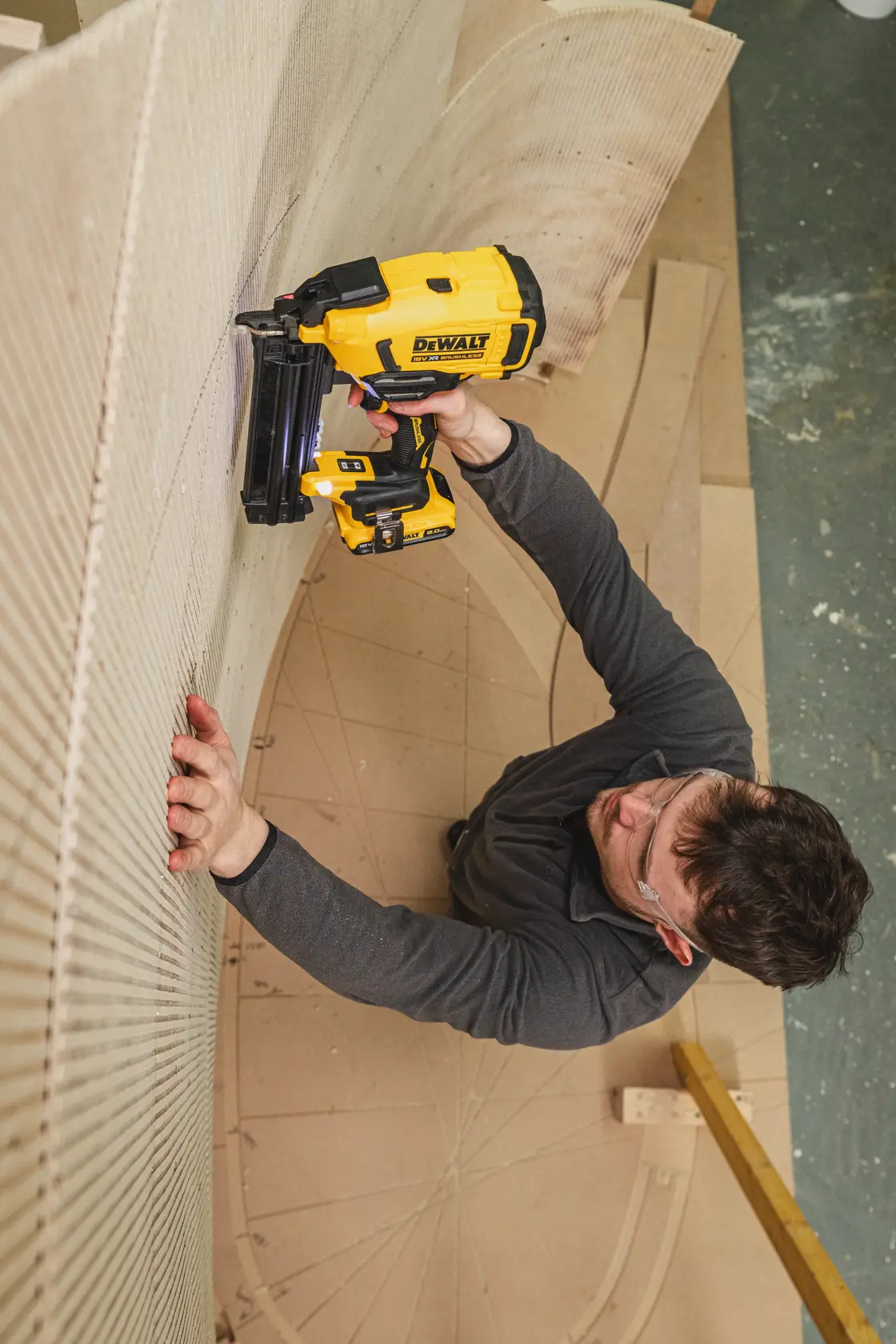 Person using DEWALT nailer DCN210SN-XJ on curved wood in workshop, wearing safety glasses and dark clothing.