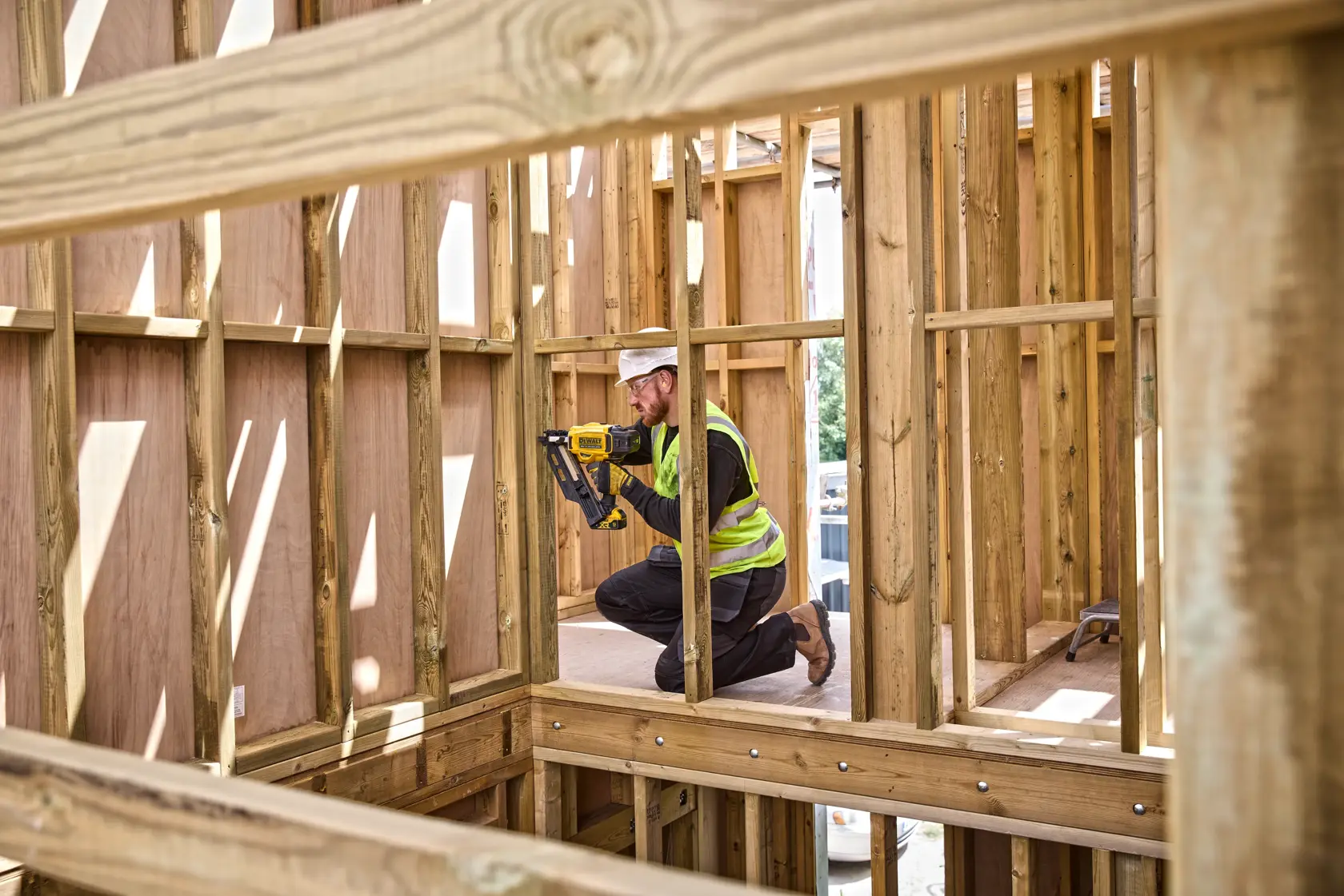 Trabajador usa clavadora DEWALT en estructura de madera. Lleva chaleco y se arrodilla en habitación sin terminar.