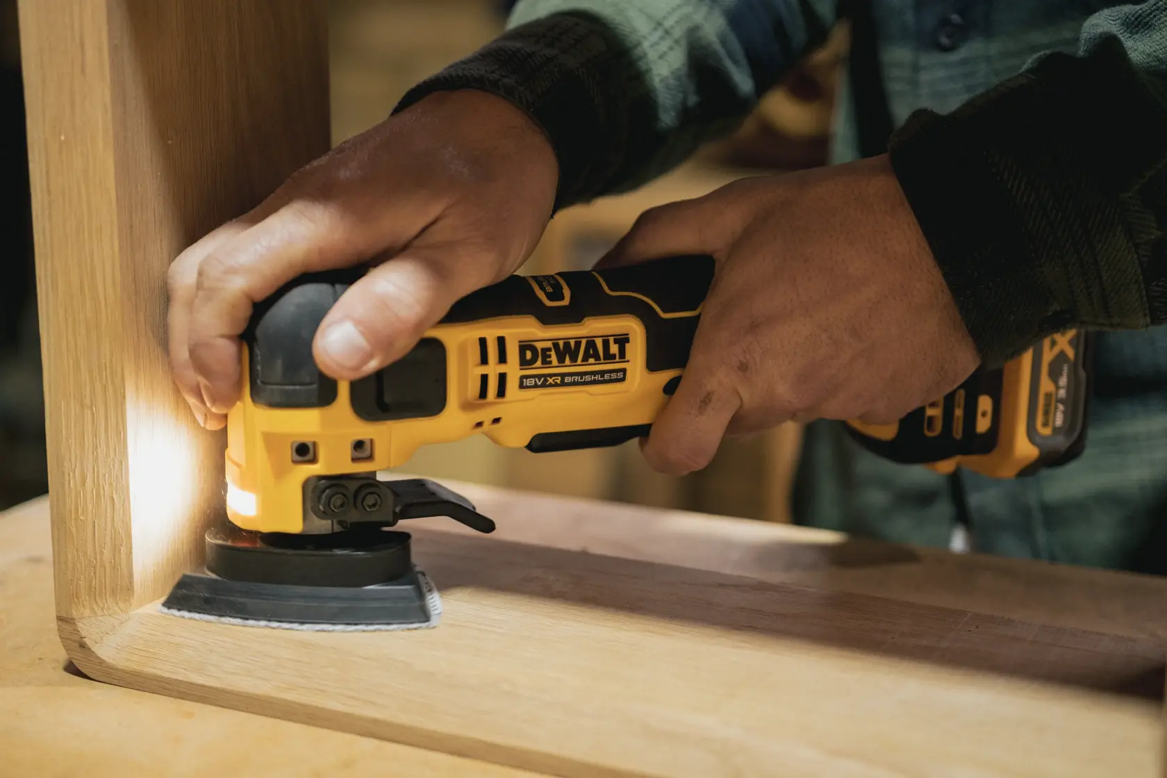 Person using a DEWALT cordless multi-tool to sand wood furniture corner indoors.