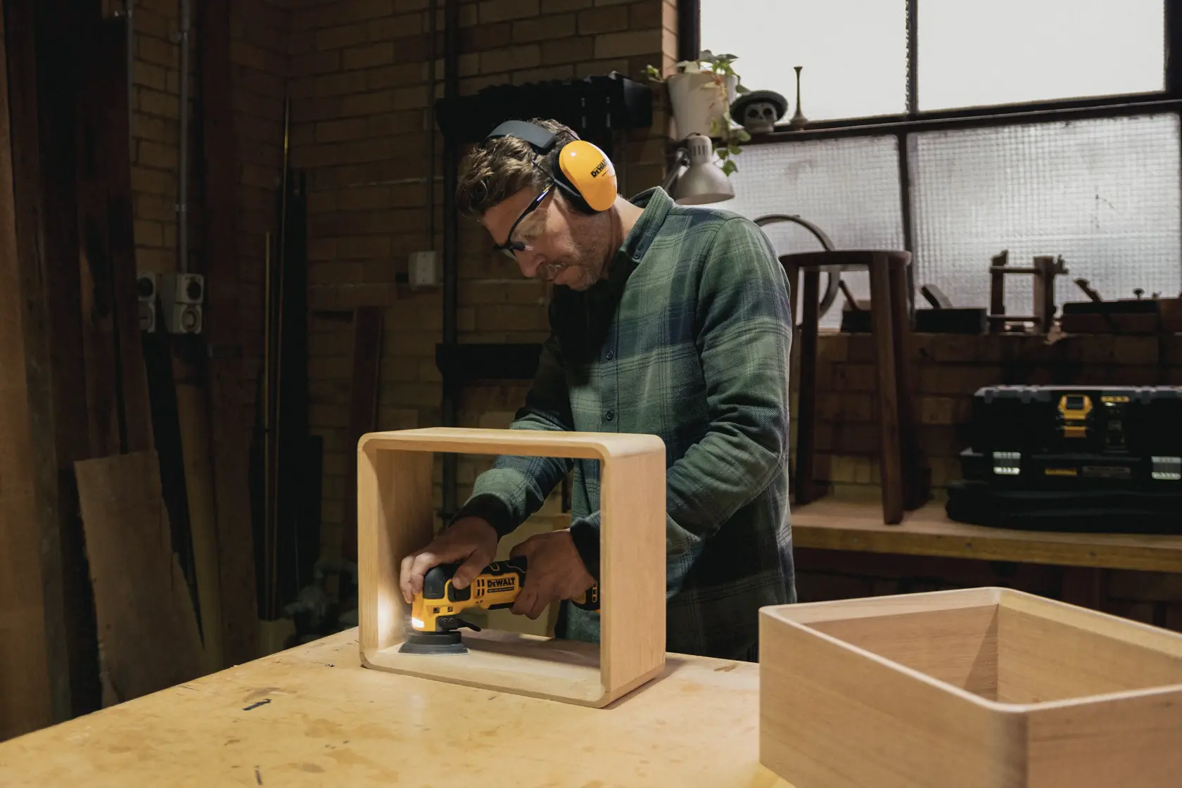 Person sanding wooden box with a DEWALT tool in a workshop wearing safety gear.