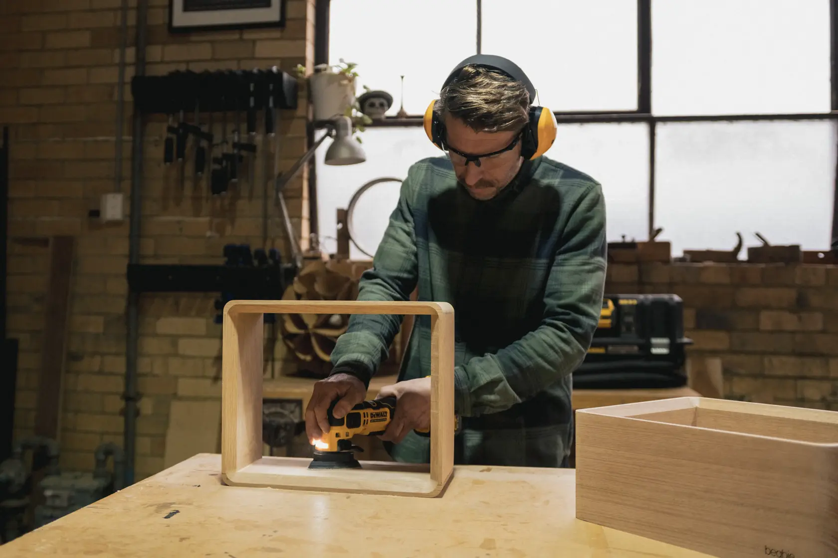 Person using a DEWALT sander on a wooden frame in a workshop, wearing ear and eye protection.