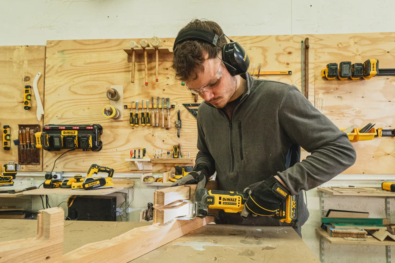 Person using a DEWALT DCS359N-XJ power tool on wood in a workshop with various tools, wearing safety gear.