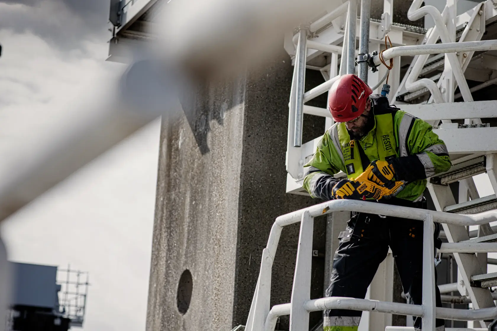 A construction worker wearing a safety helmet and high-visibility jacket is using a DEWALT power tool while standing on an industrial metal platform. The worker is secured with a safety harness.