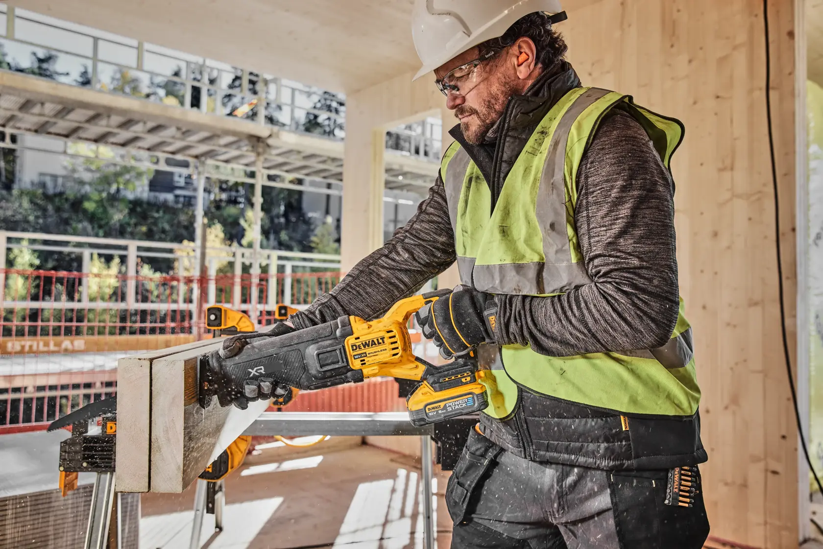A person wearing a safety vest and hard hat is using a DEWALT cordless reciprocating saw (model DCS386H2T) to cut a piece of wood on a construction site.