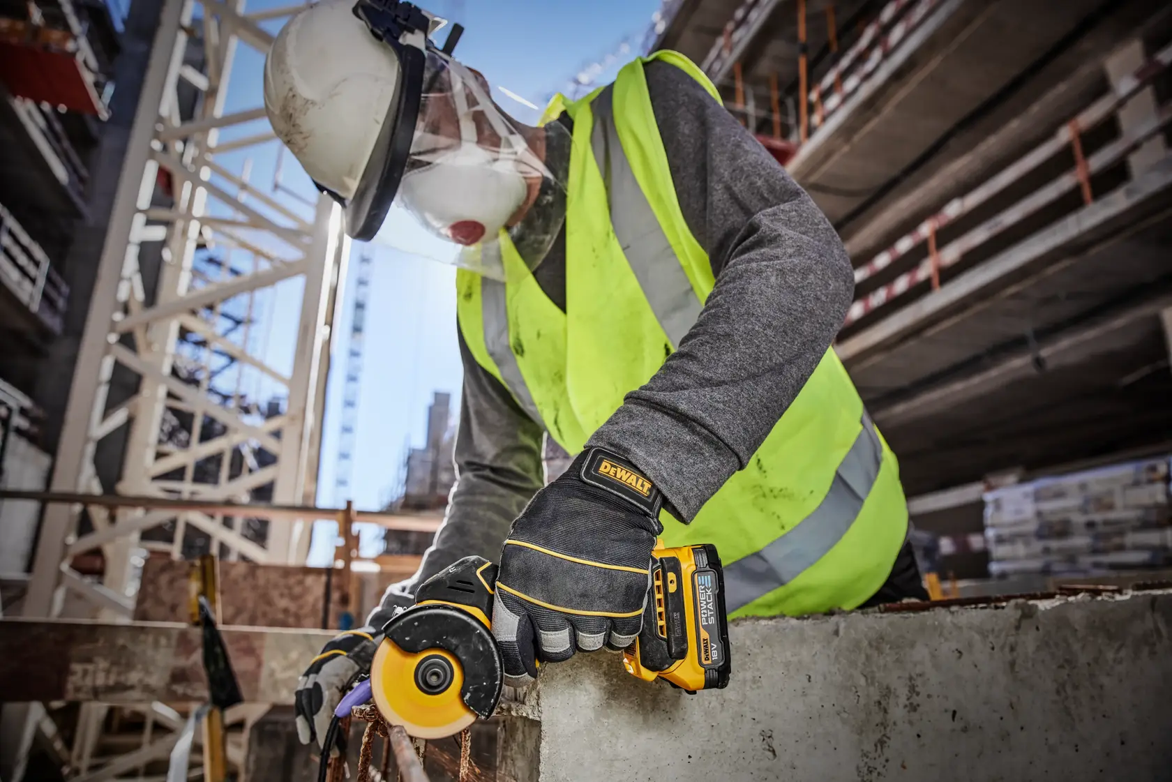 Un trabajador de la construcción con casco de seguridad, visera facial, chaleco reflectante y guantes utiliza una herramienta inalámbrica DEWALT DCS438N para cortar concreto en un sitio de construcción exterior.