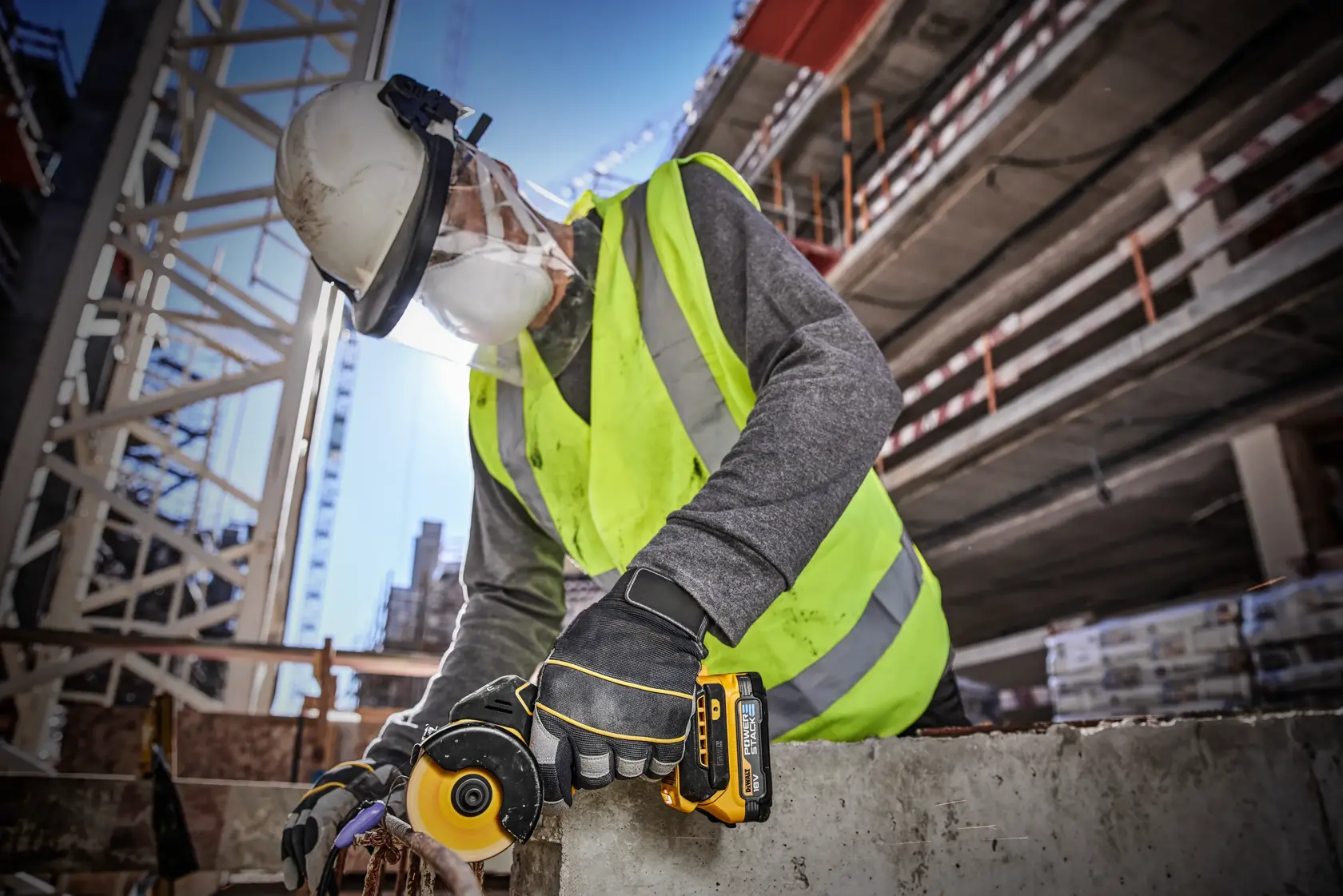 Un trabajador de la construcción lleva un casco de seguridad, chaleco de alta visibilidad, guantes y careta protectora, y utiliza una herramienta inalámbrica DEWALT DCS438N para cortar o pulir hormigón en una obra.