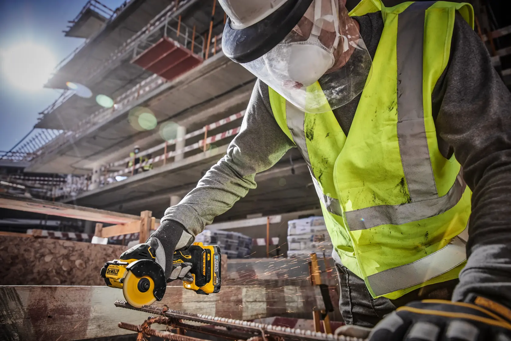 Un trabajador de la construcción con chaleco de alta visibilidad, guantes protectores, mascarilla y casco utiliza una herramienta eléctrica inalámbrica DEWALT DCS438N para cortar barras de metal en una obra con una estructura parcialmente construida al fondo.