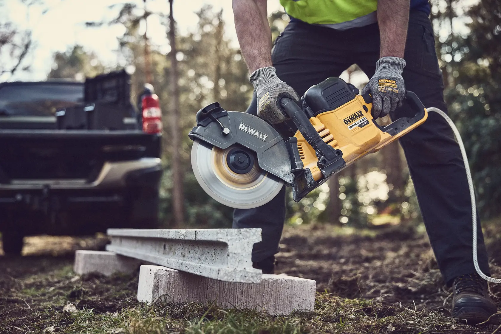 Persona corta concreto al aire libre con una sierra DEWALT. Guantes protectores y camioneta al fondo.