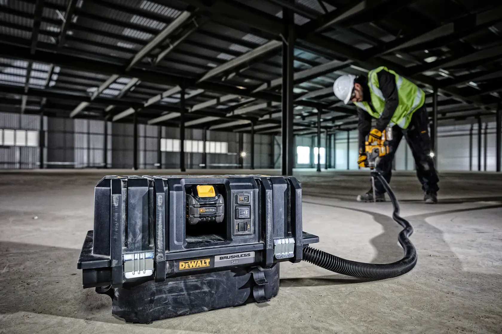 Aspiradora DEWALT DCV586MT2 en piso de obra, trabajador perfora al fondo usando equipo de seguridad.