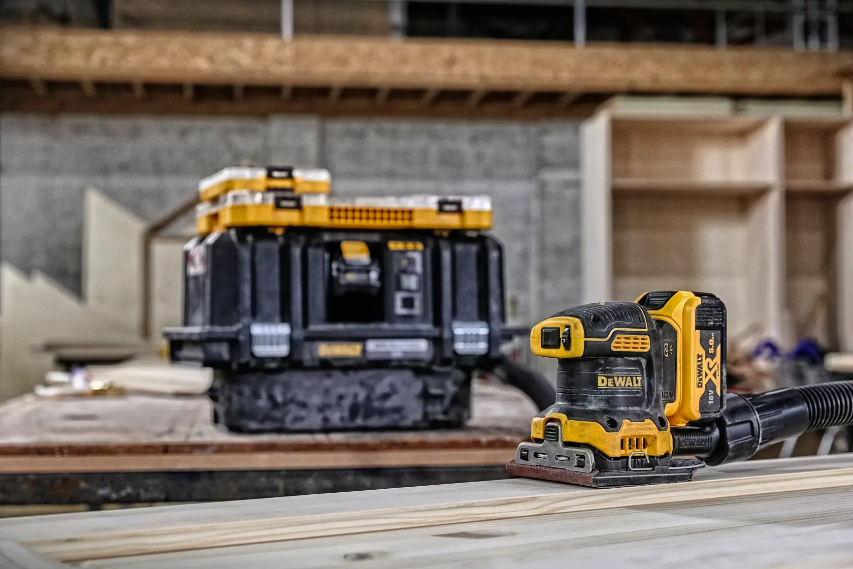 Lijadora inalámbrica DEWALT sobre madera, caja de almacenamiento al fondo en taller.