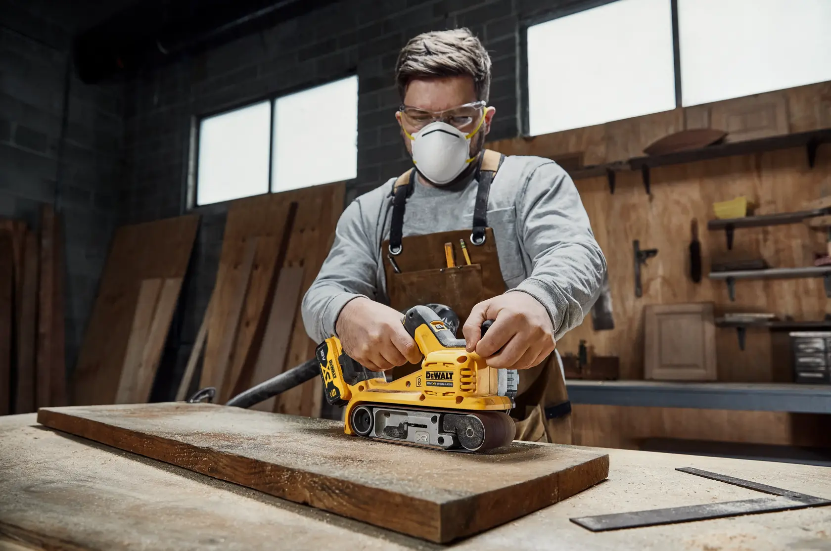 A person in a workshop using a DEWALT DCW220P2 cordless belt sander to smooth a wooden plank on a table. The person is wearing a brown apron and safety equipment, and the workspace has wooden boards and tools visible in the background.