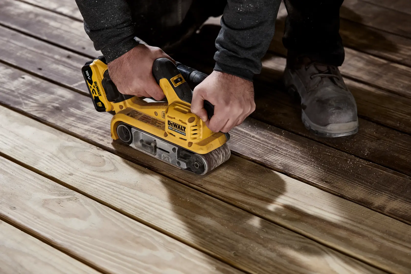 A person using a yellow DEWALT cordless belt sander (model DCW220P2) to sand wooden floorboards.