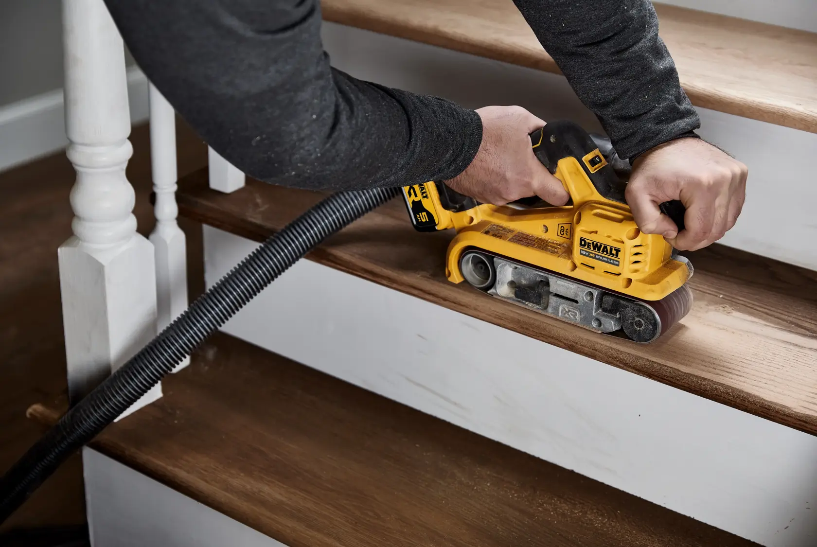 A person is using a DEWALT DCW220P2 yellow cordless belt sander to sand a wooden staircase step. A vacuum hose is attached to the tool for dust extraction.