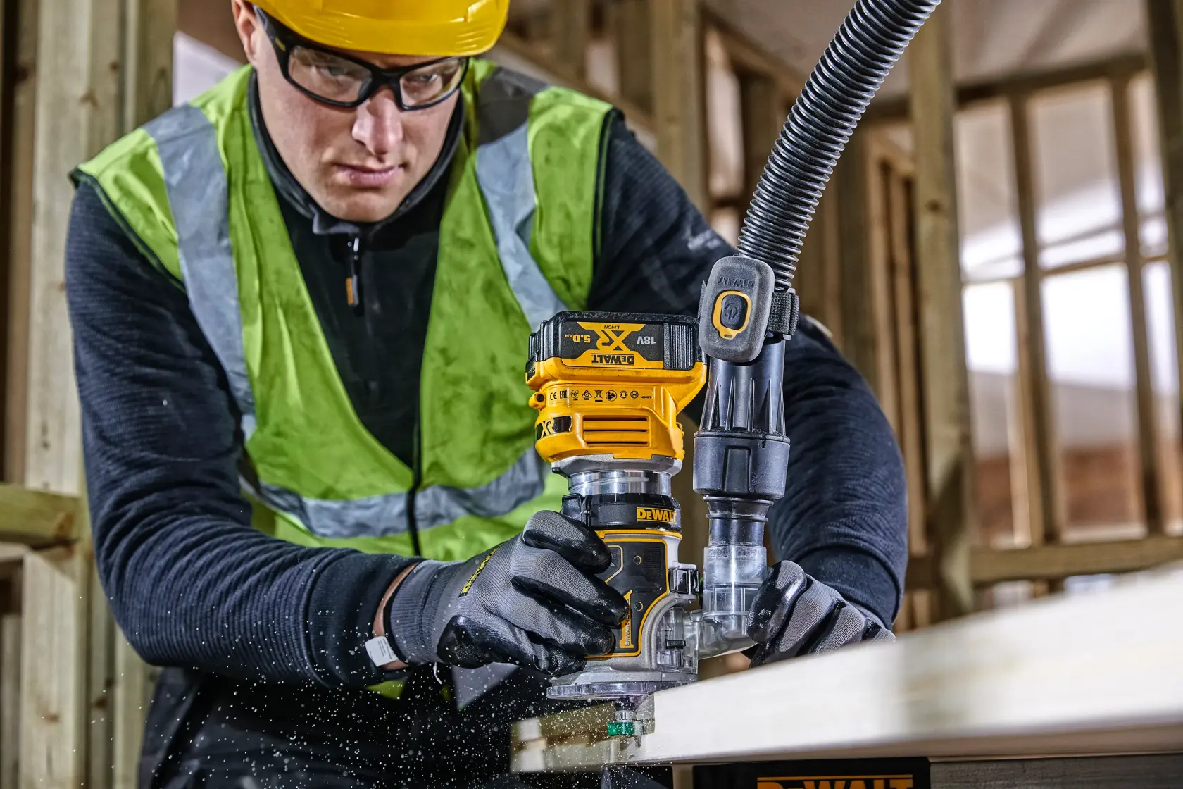 A person uses a DEWALT DCW604 router to shape wood in a construction setting, wearing safety gear and gloves.
