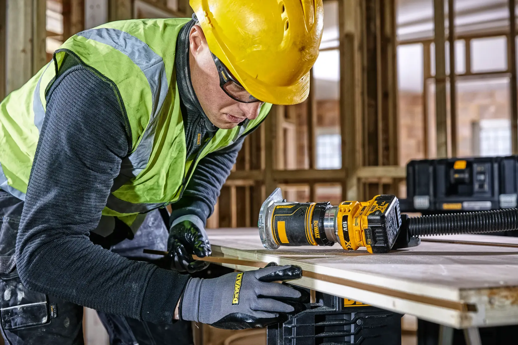 Worker uses DEWALT DCW604 router tool on wood in a construction site, wearing gloves, helmet, and safety vest.