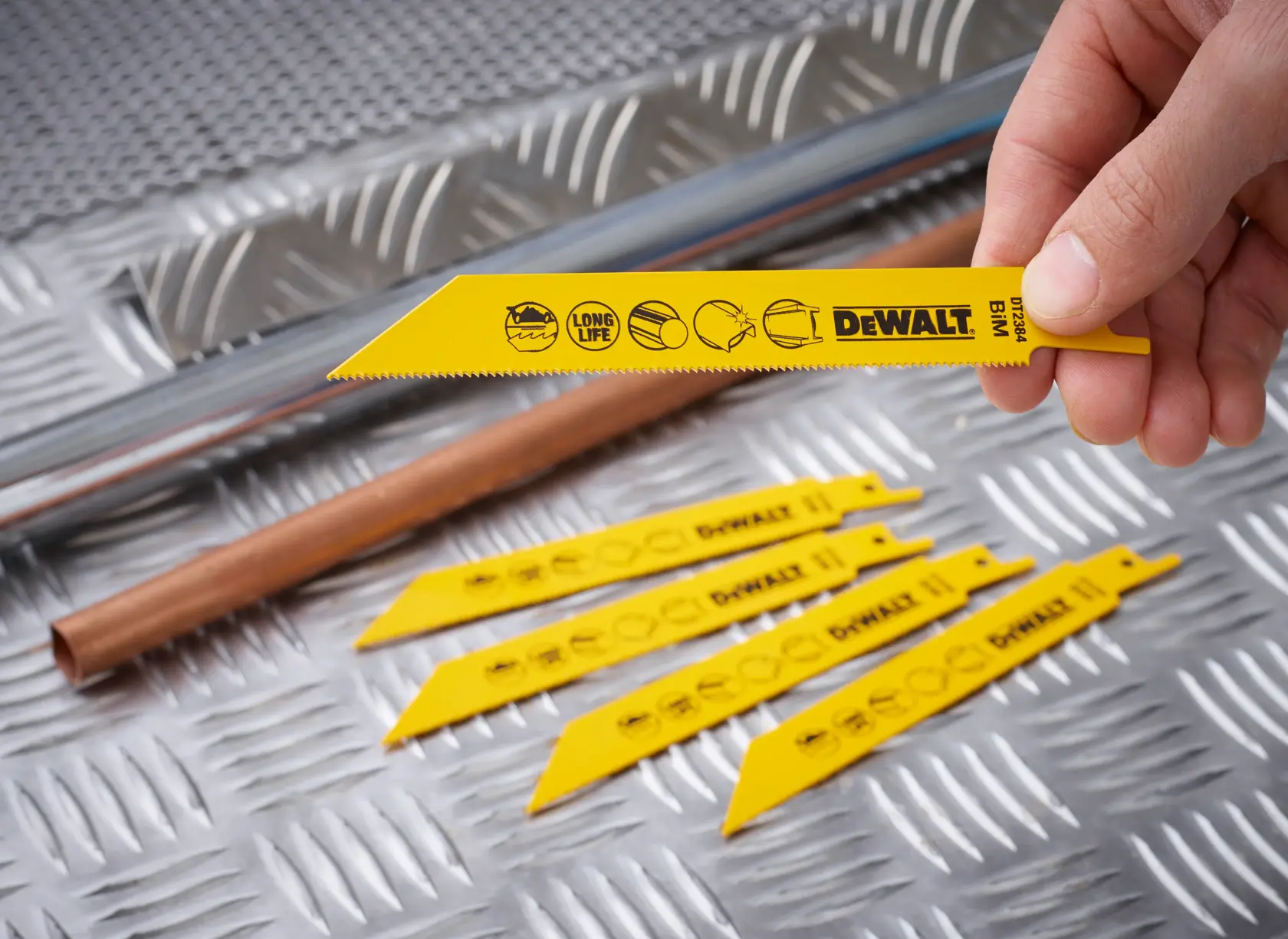 A hand holds a yellow DEWALT DT2384 saw blade with others lying on metal sheet beside metal pipes.