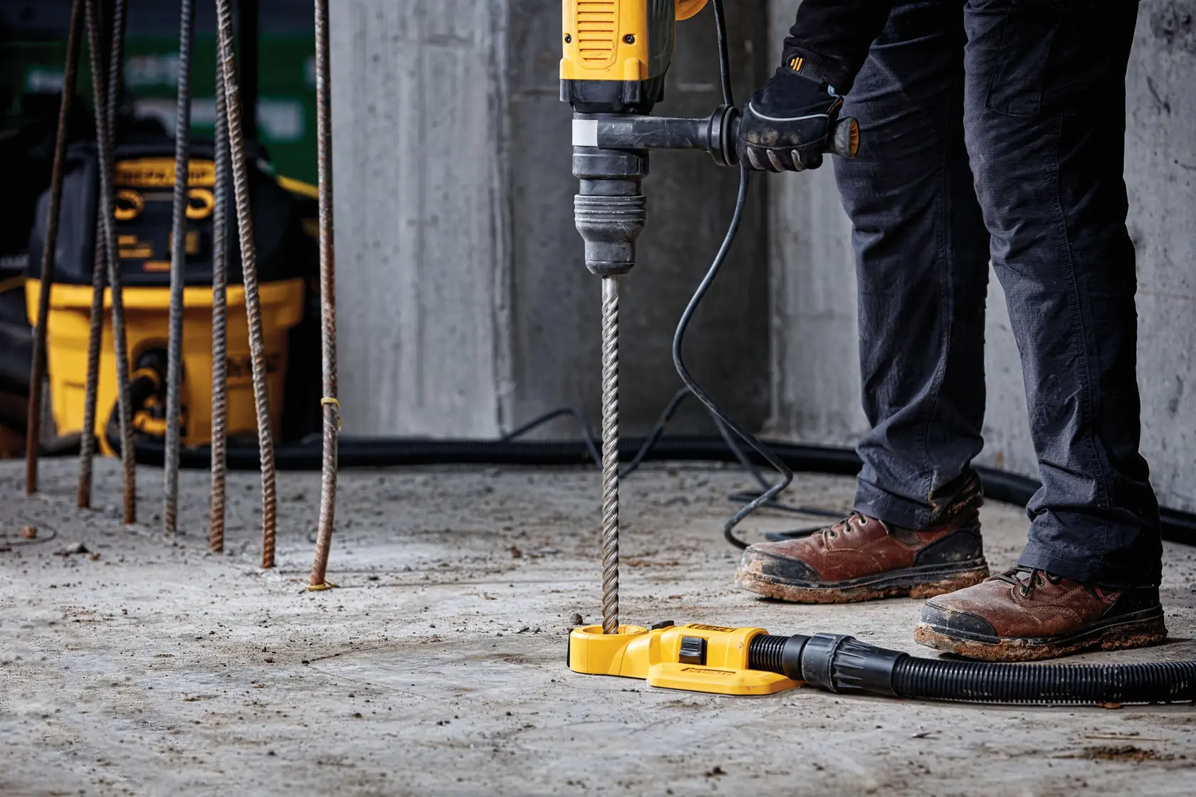 Person using a DEWALT hammer drill with dust extraction accessory on a concrete floor at a construction site.
