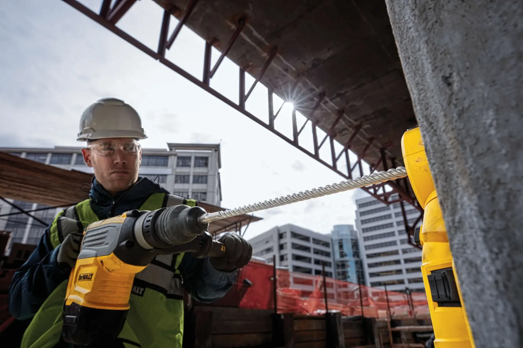 Worker uses a DEWALT drill with a long bit at a construction site with buildings in the background.