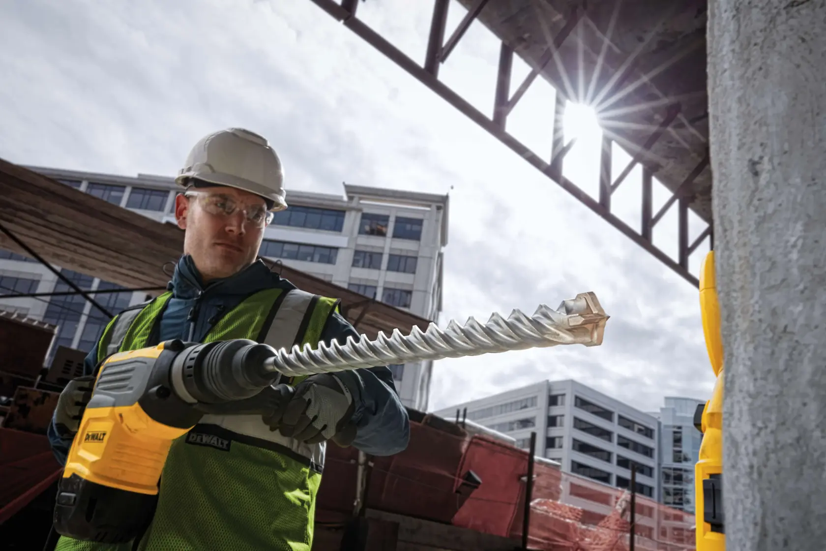 Person holding a DEWALT DT60846 rotary hammer drill with spiral bit at a construction site, buildings in the background.