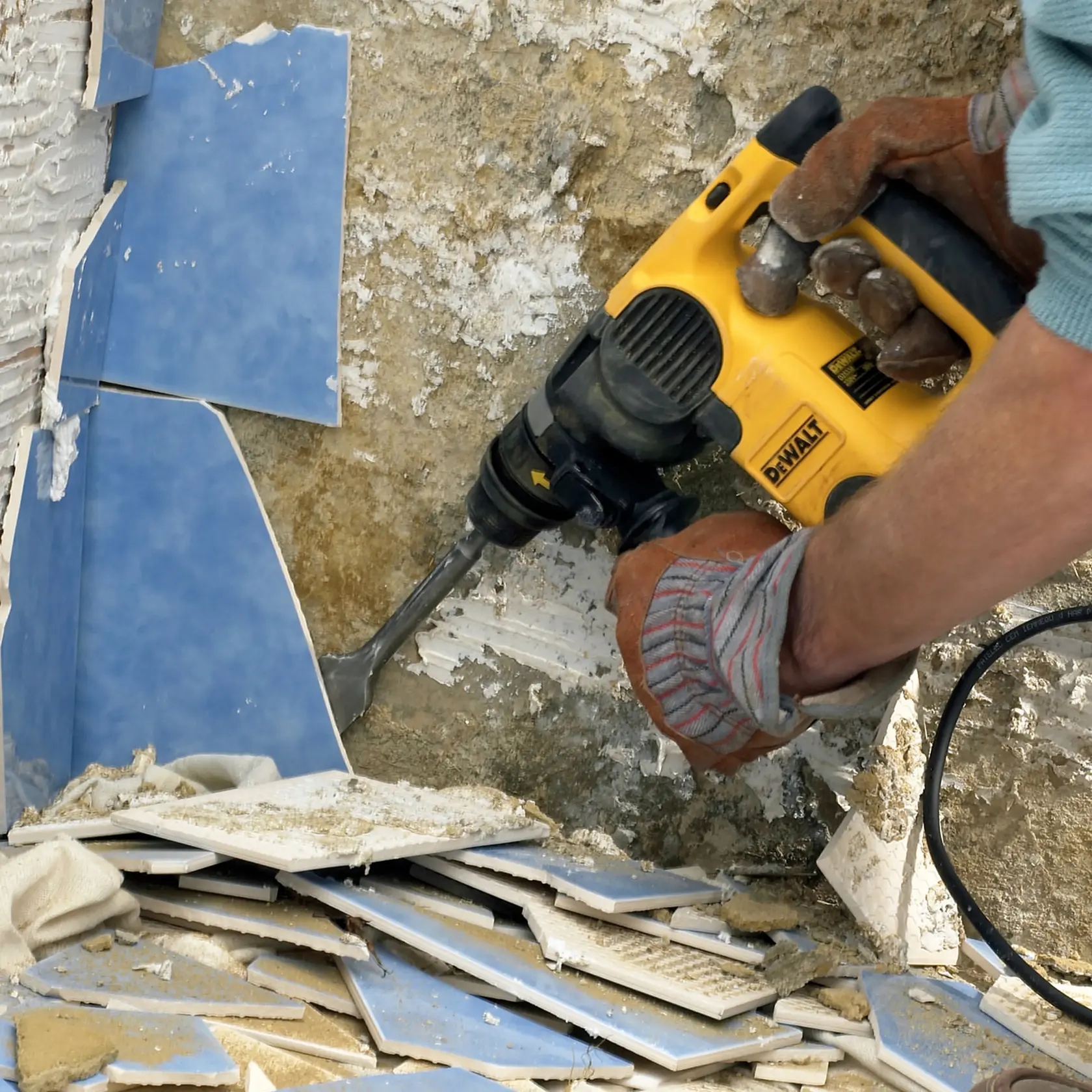 Photo of DEWALT DT6803 chisel in use, removing blue tiles from wall, yellow tool visible, gloves on hands.