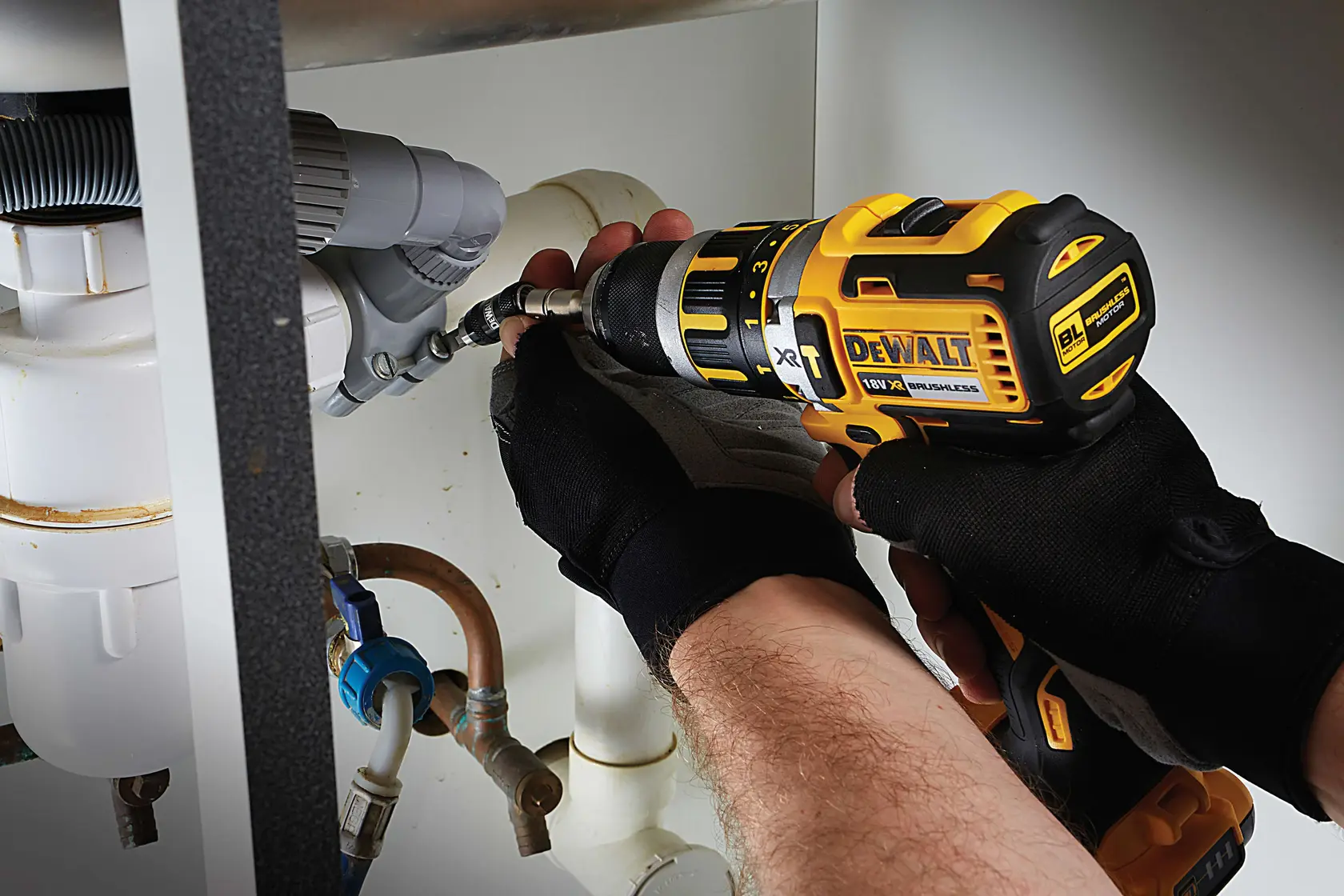 Photo of DEWALT DT70518T drill in use by gloved hands, working on plumbing under a sink.