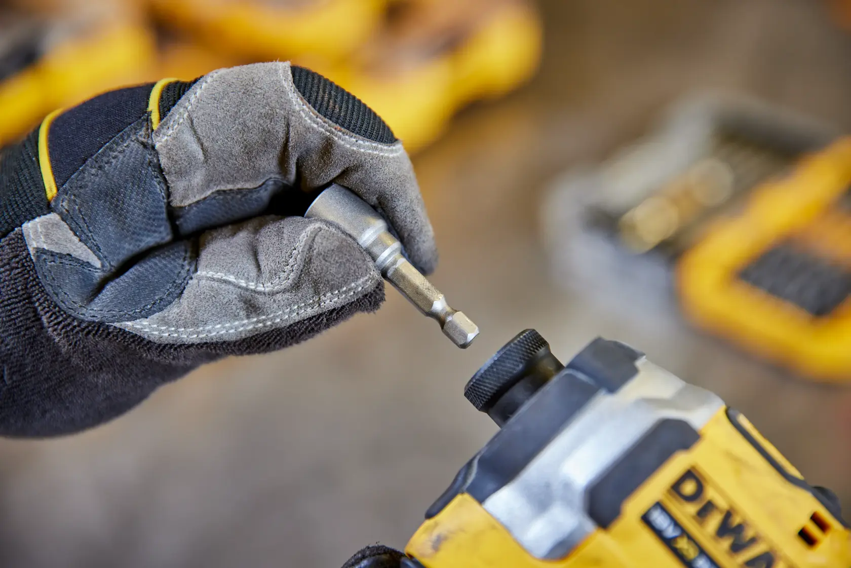 Gloved hand inserts a DEWALT bit into a yellow power tool. Tool kits are blurred in the background.
