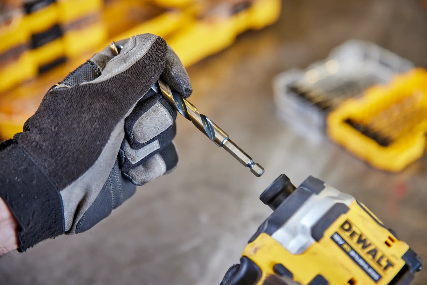 Gloved hand holding drill bit near DEWALT tool with yellow storage case in background.