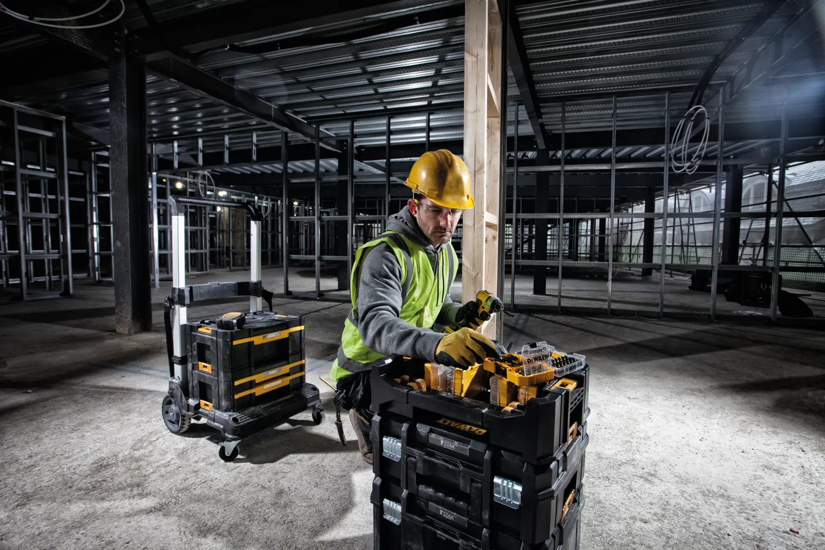 Worker in safety gear using DEWALT toolboxes at a construction site with metal framing and unfinished walls.