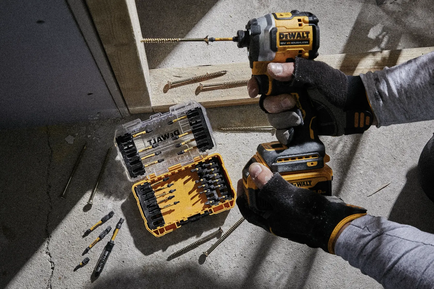 Person uses DEWALT drill near open bit set case and screws on concrete floor at a construction site.