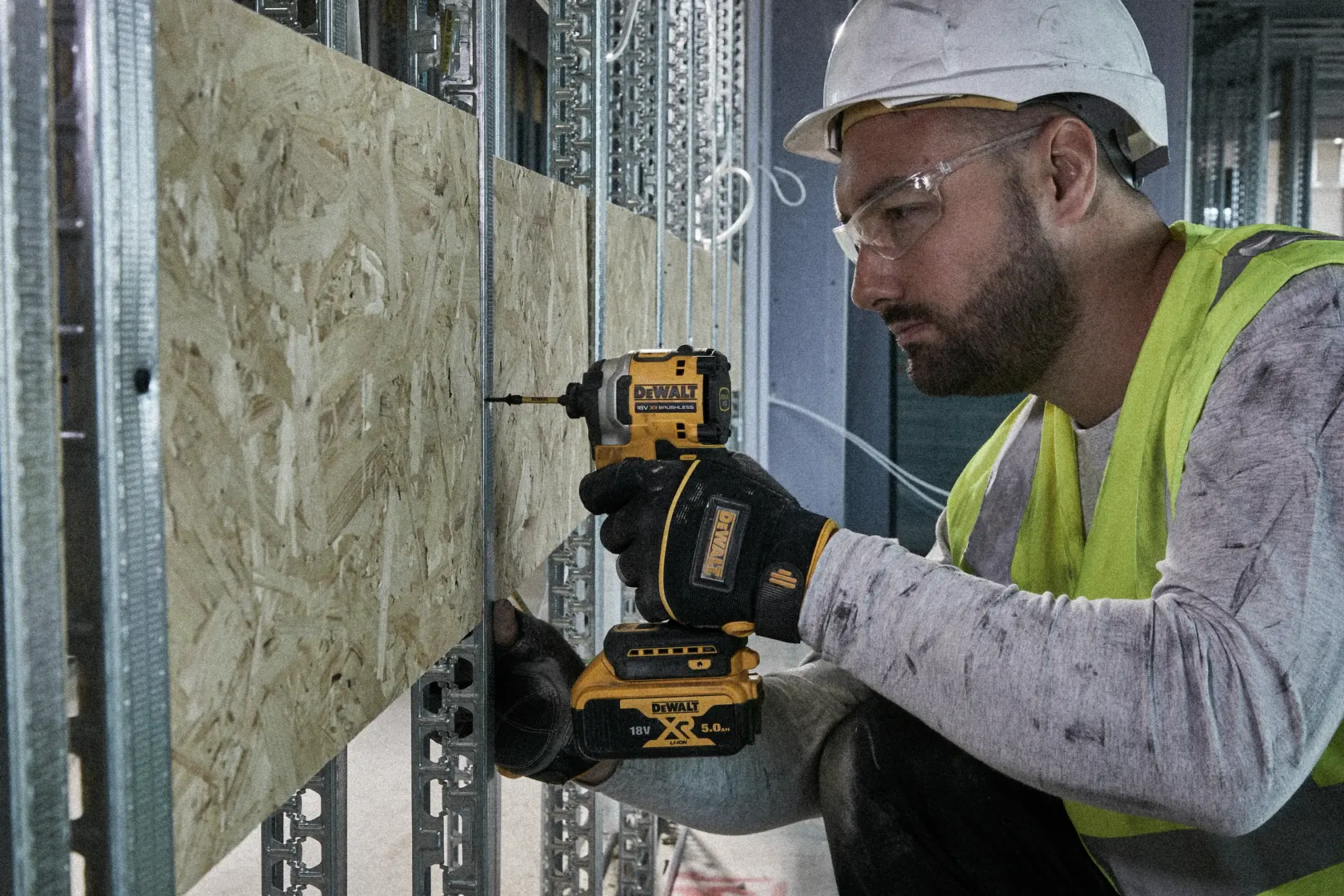 Person using a DEWALT drill on a wooden panel at a construction site, wearing safety gear and gloves.