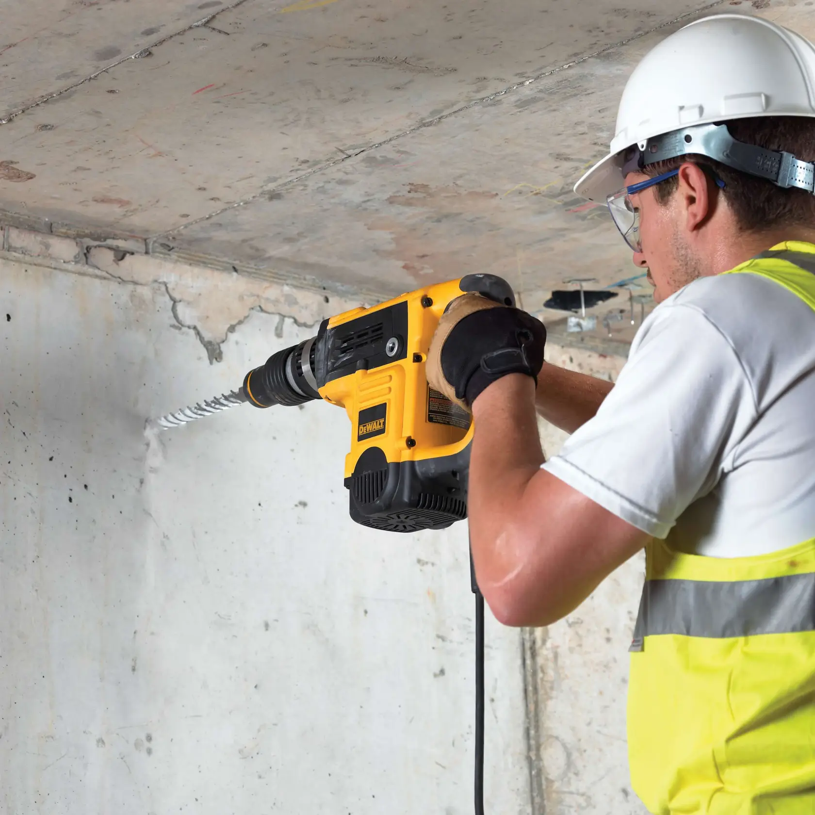 Worker drills into concrete wall with DEWALT power tool. Safety gear worn, including helmet and gloves.