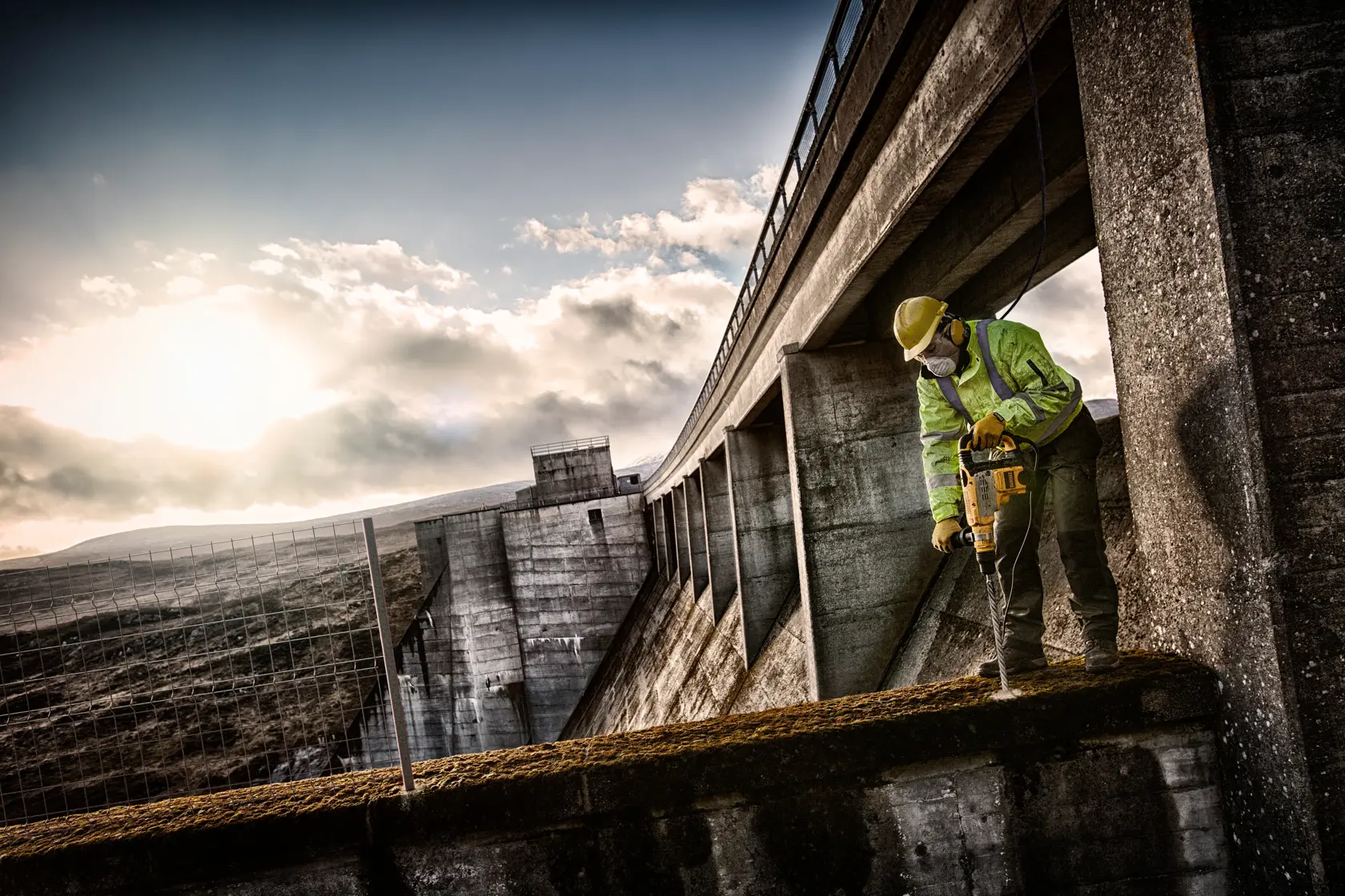 Worker in safety gear using a DEWALT drill on concrete structure outdoors near a dam, with dramatic sky in background.