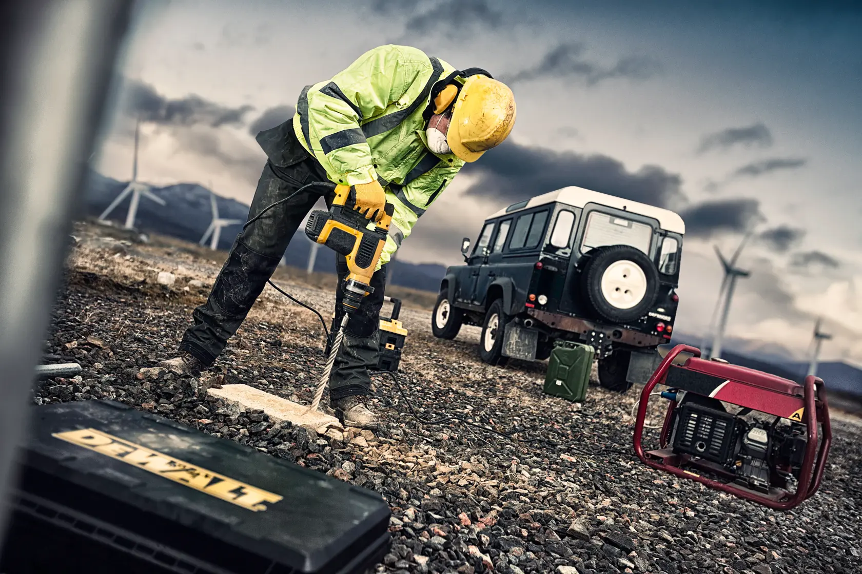 Worker uses DEWALT drill on rocky ground, with toolbox, SUV, and wind turbines in the background outdoors.