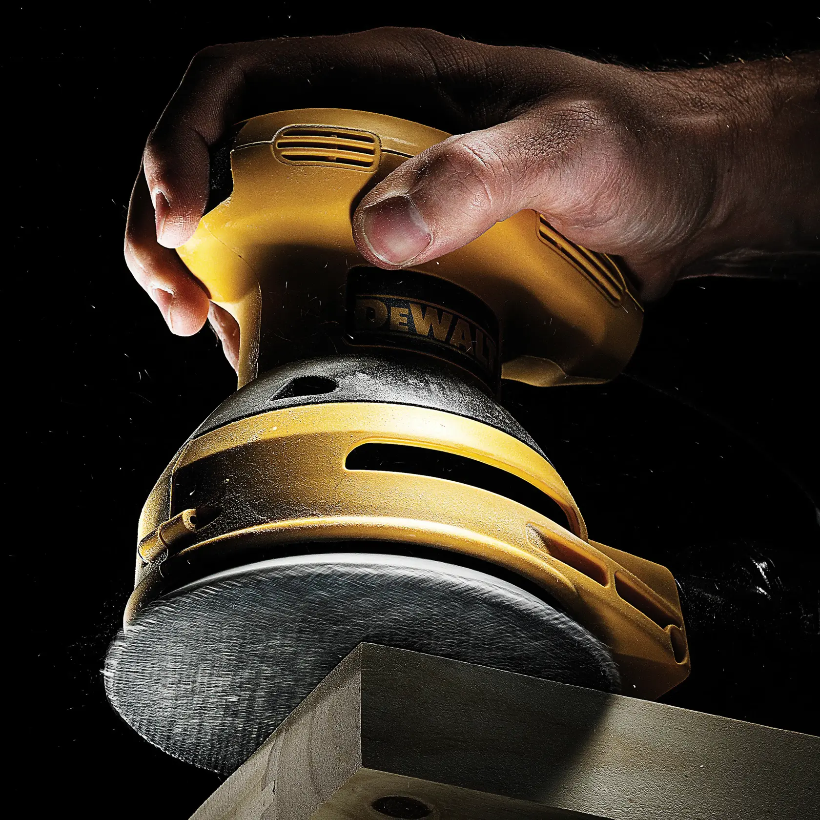 Hand holding DEWALT sander, smoothing wood surface. Close-up view, dust visible, black background.