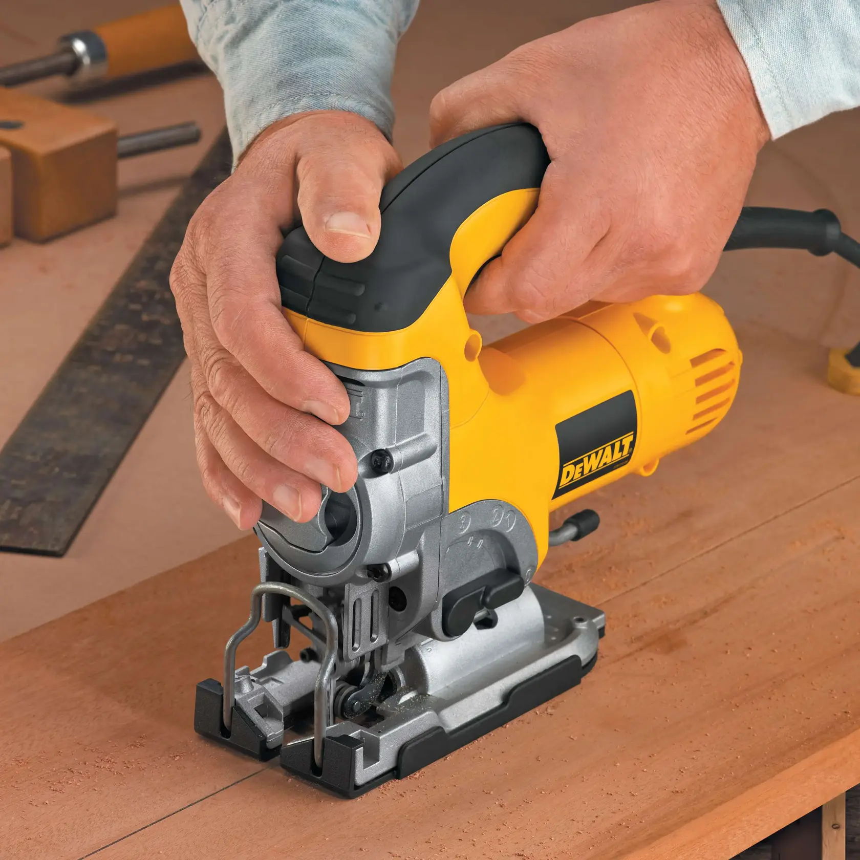Manos usan una sierra de calar DEWALT para cortar madera en mesa de trabajo. Hay otras herramientas detrás.