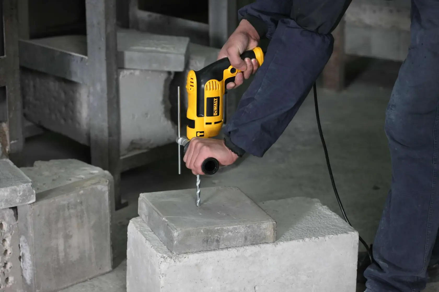 A person uses a yellow DEWALT electric drill to drill into a concrete block. The person is holding the drill with both hands for stability in an industrial setting.