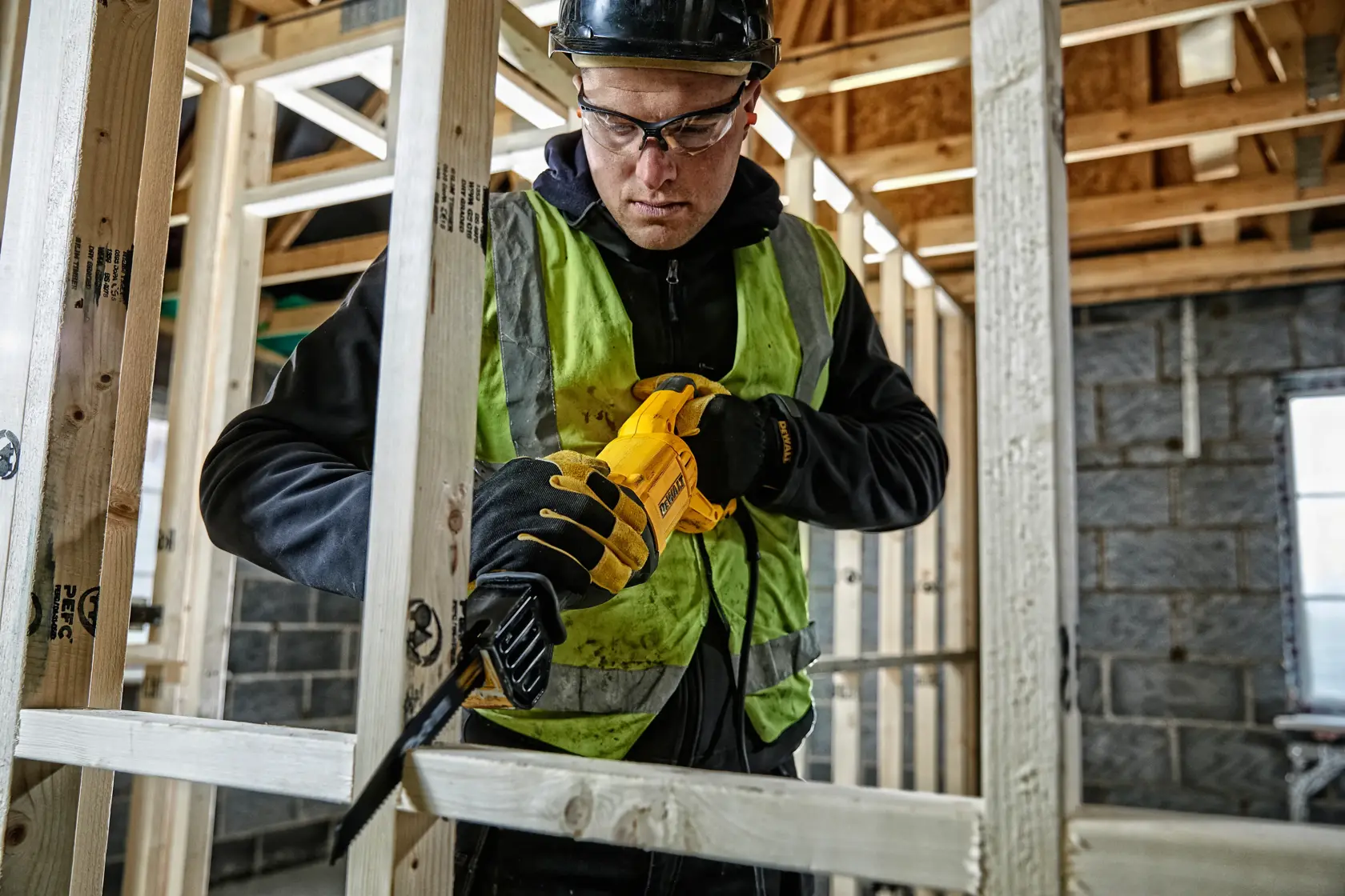 Trabajador corta madera con sierra amarilla DEWALT en obra, lleva chaleco y casco de seguridad.